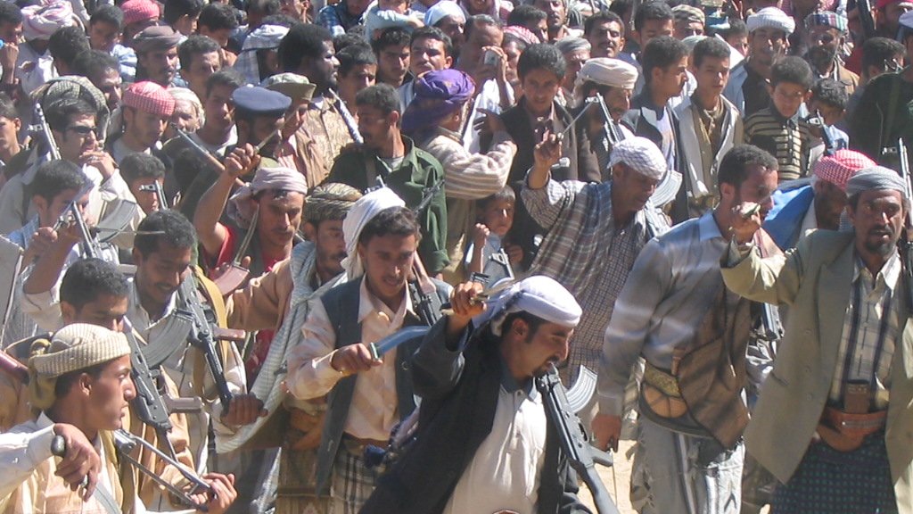 L'image montre un groupe de personnes en pleine célébration ou manifestation culturelle. Les hommes portent des vêtements traditionnels, y compris des chapeaux et des écharpes. Certains sont armés avec des fusils, ce qui pourrait indiquer un contexte de fête ou de rite traditionnel dans une région où la possession d'armes est courante. L'atmosphère semble dynamique et vivante, avec les participants se déplaçant en rythme, probablement en dansant ou en chantant.