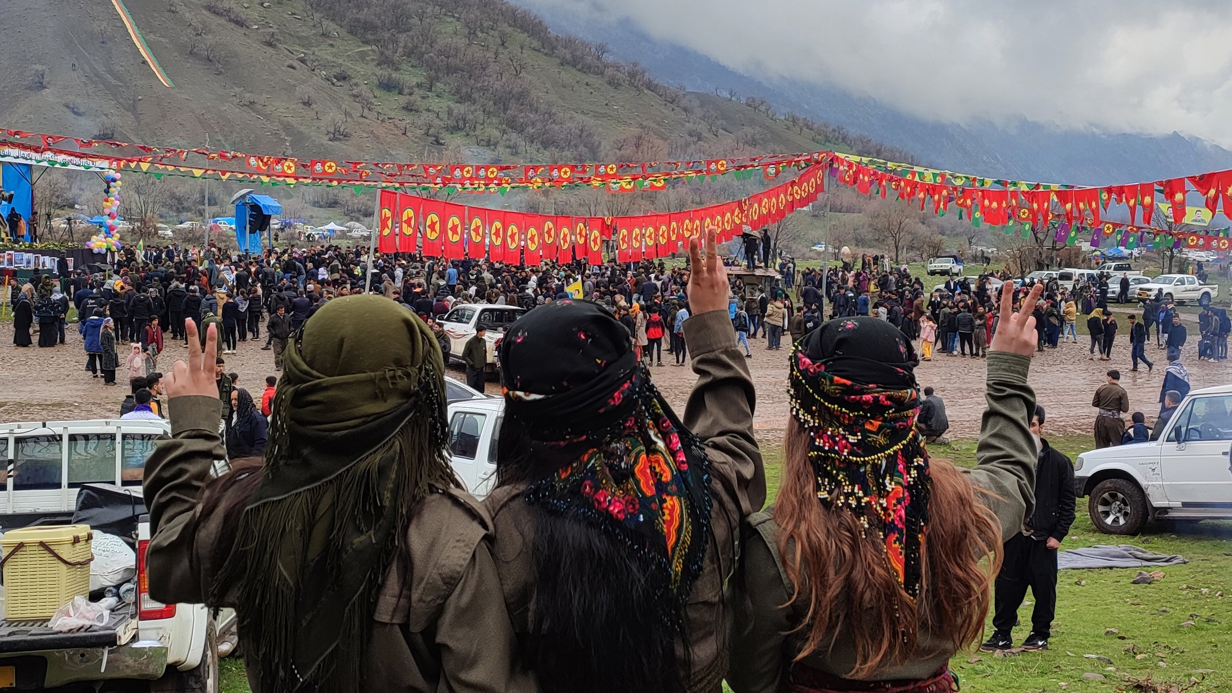 L'image montre un groupe de trois personnes vues de dos, faisant un signe de paix avec leurs mains. Elles portent des vêtements traditionnels et des foulards colorés. En arrière-plan, on peut voir une foule rassemblée pour un événement, avec des banderoles rouges décorées de symboles. Le paysage environnant est montagnard et sous un ciel nuageux. L'atmosphère semble festive et solennelle.
