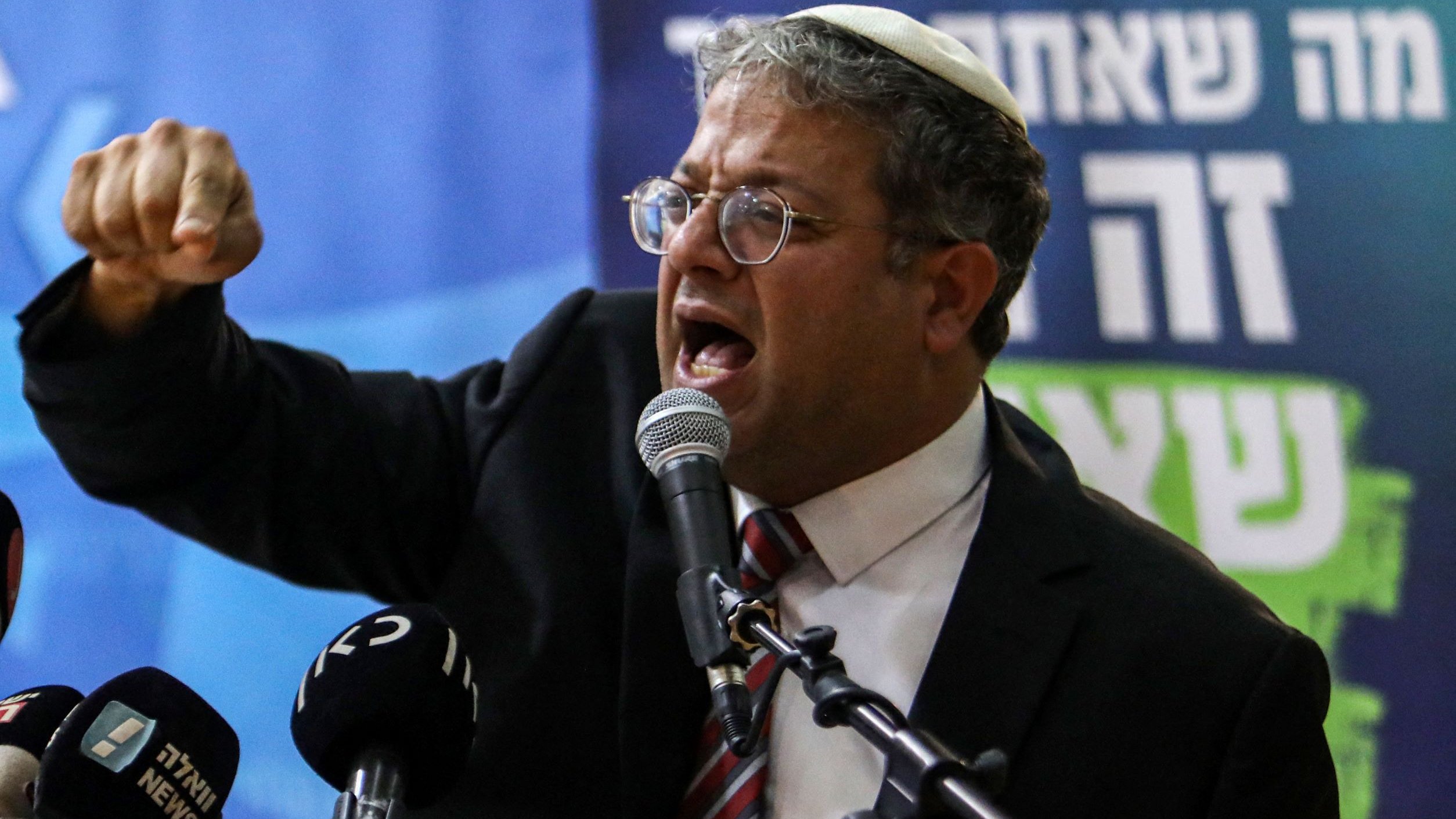 L'image montre un homme portant des lunettes et une kippa, en train de s'exprimer devant un micro. Il semble passionné par ses propos, faisant un geste avec son bras droit tendu et son poing fermé. Il porte un costume sombre avec une cravate rayée. Devant lui, plusieurs micros de différentes chaînes de presse sont visibles. L'arrière-plan montre une bannière avec du texte en hébreu.