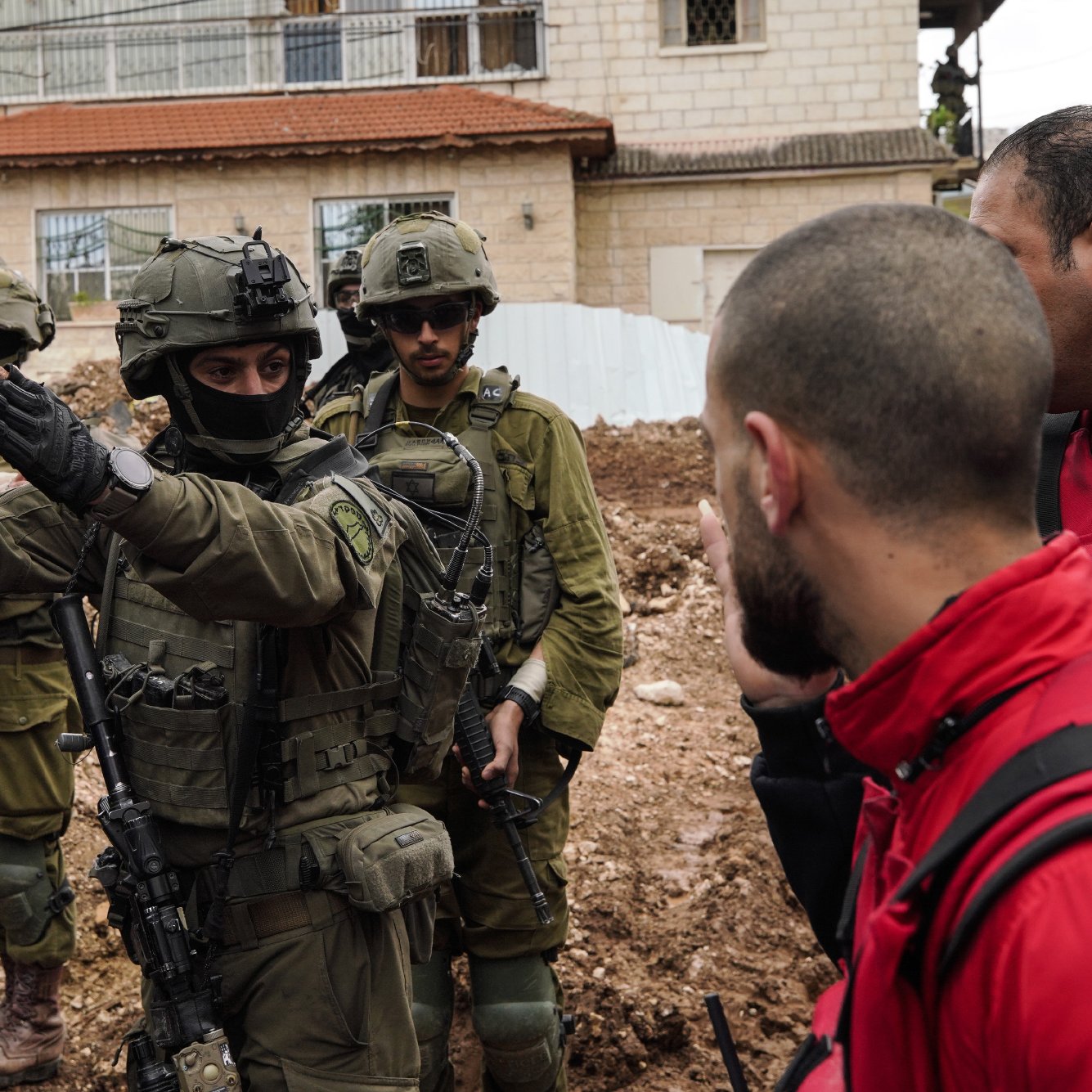 Nell'immagine ci sono diversi uomini in un contesto urbano, alcune persone indossano giacche protettive e una di loro è in uniforme militare con un'arma. Sembra esserci una discussione in corso tra le persone. Il terreno attorno a loro è fangoso e c'è un edificio sullo sfondo.