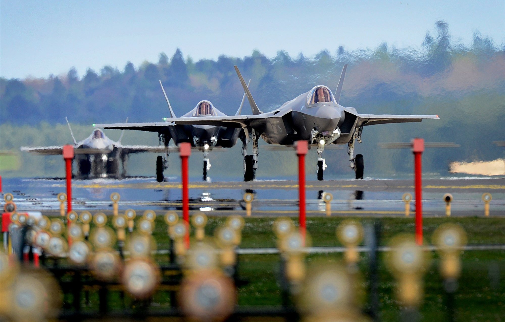 L'image montre plusieurs avions de chasse F-35 alignés sur une piste d'atterrissage. Au premier plan, on peut voir des lumières et des repères au sol, tandis que les avions sont prêts au décollage. En arrière-plan, on aperçoit une forêt verdoyante sous un ciel clair. L'ambiance général semble être celle d'une base aérienne, mettant en avant la puissance et la technologie militaire moderne.