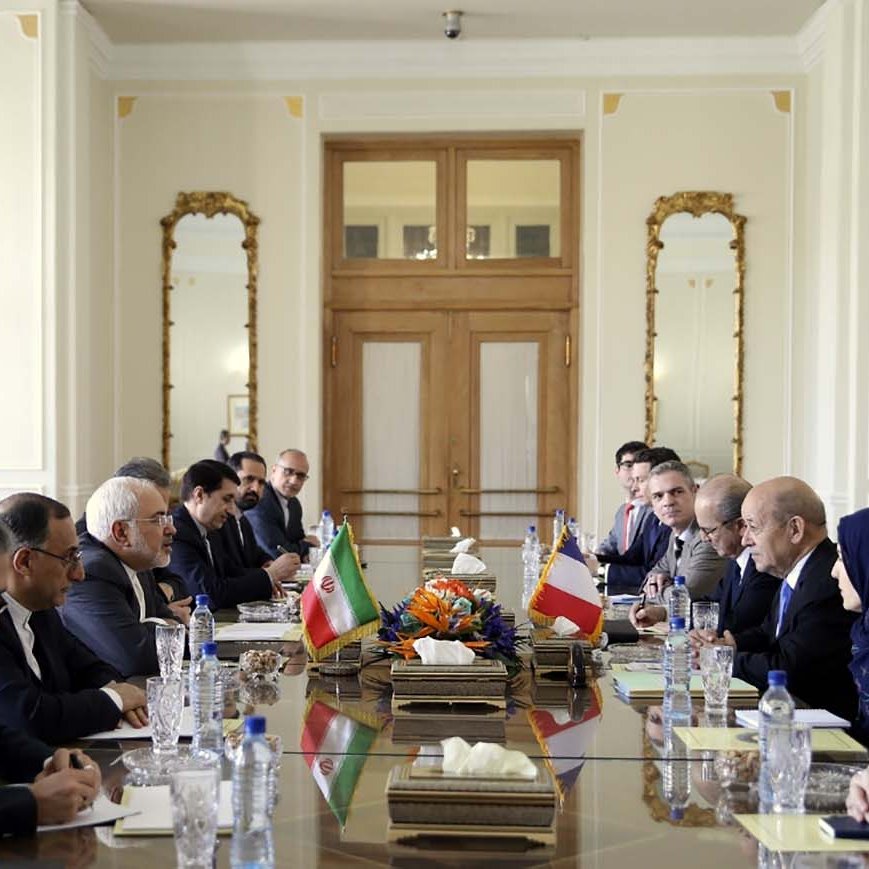 تظهر في الصورة قاعة اجتماعات تجمع وفدين من رجال السياسة. يجلس الأعضاء على طاولة طويلة مرتبة بشكل منظم، حيث توجد أعلام إيران وفرنسا على الطاولة. يتواجد في الجانبين رجال ونساء يرتدون ملابس رسمية، ويبدو أن النقاشات جادة ومتبادلة. تعكس الأجواء في الغرفة رسمية واحترافية، مع تفاصيل معمارية متقنة في المكان.