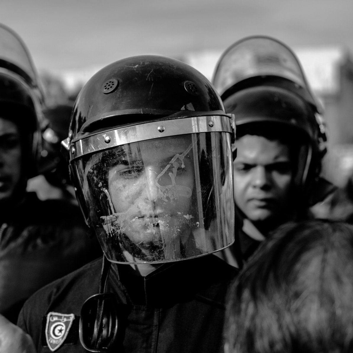 L'image montre un groupe de personnes, probablement des agents des forces de l'ordre, entourés par une foule. L'un des agents est au premier plan, portant un casque et un bouclier facial, ce qui indique qu'il est en position de maintien de l'ordre. Les visages des autres personnes en arrière-plan sont également visibles, exprimant une certaine tension. L'image est en noir et blanc, ce qui accentue le sérieux de la situation. L'atmosphère semble chargée, avec un sentiment de confrontation possible.