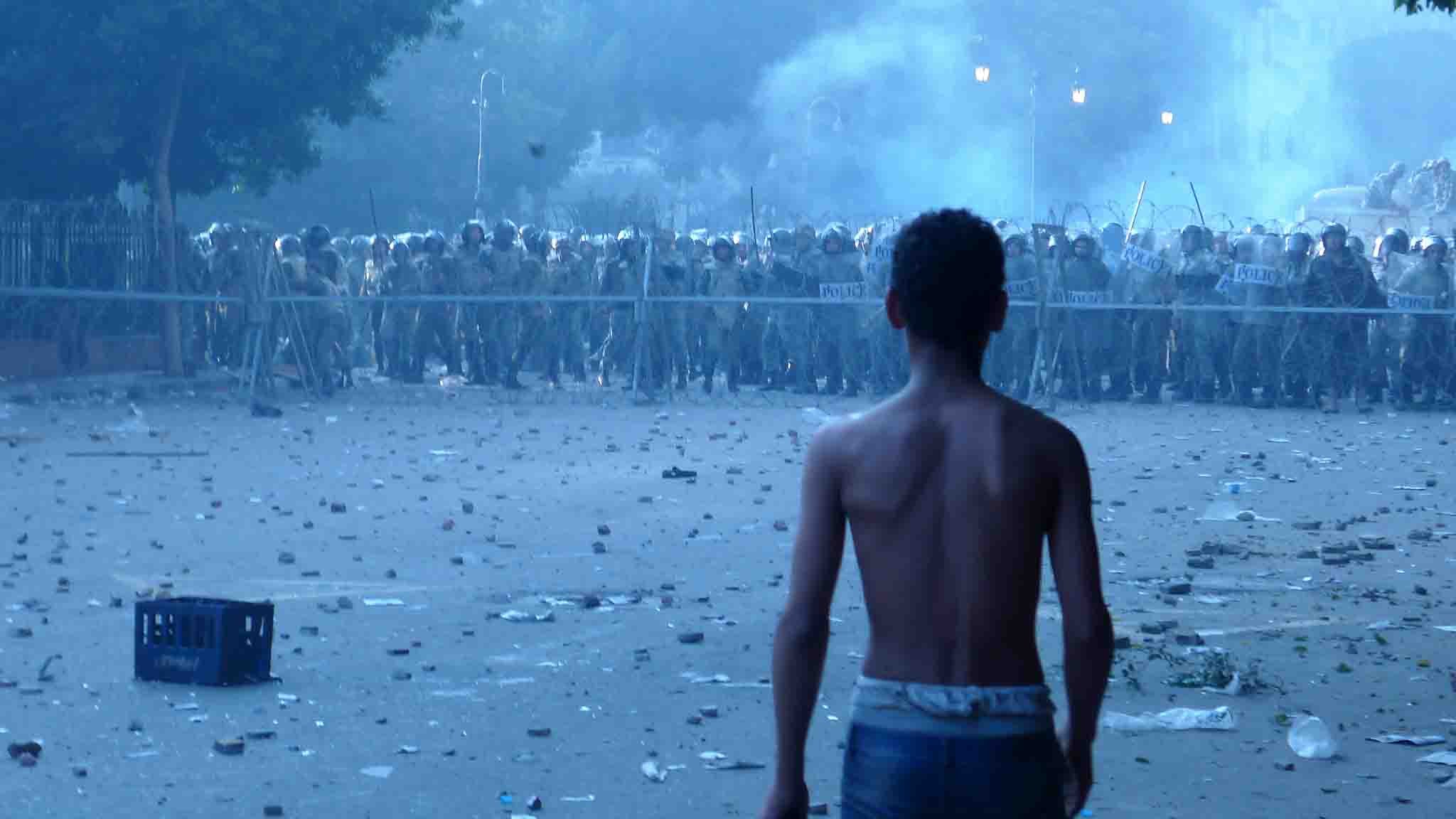 تصویر نشان‌دهنده یک صحنه تنش‌زا است. در جلو، یک پسر جوان با تنه برهنه به سمت گروهی از نیروهای انتظامی که در فاصله‌ای دورتر ایستاده‌اند، نگاه می‌کند. این نیروها در لباس‌های نظامی و با سپرهایی در دست، آماده مواجهه هستند. زمین در اطراف پر از زباله و بقایای ناآرامی‌هاست، و بخاری یا دودی در هوا وجود دارد که جو پرتنش‌تری را ایجاد می‌کند. فضای کلی تصویر احساس اضطراب و کشمکش را منتقل می‌کند.