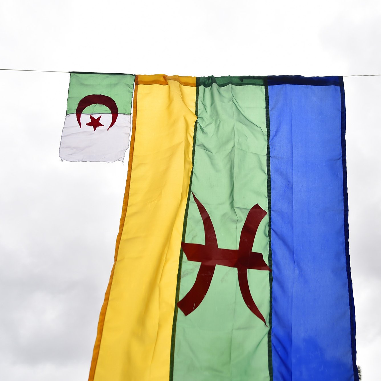 L'image montre plusieurs drapeaux suspendus. Au centre, il y a un grand drapeau de couleur verte avec un symbole rouge au centre, probablement représentant le mouvement amazigh. De chaque côté, il y a de plus petits drapeaux, qui semblent être le drapeau algérien, avec les couleurs verte et blanche et le croissant et l'étoile rouges. L'ensemble est accroché sous un ciel nuageux, créant une ambiance festive ou symbolique.