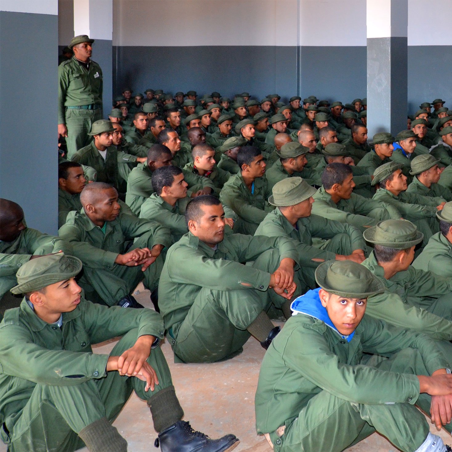 L'image montre un grand groupe de soldats assis dans une salle. Ils portent des uniformes verts et des bottes noires, et sont alignés en rangées sur le sol. Un instructeur, également en uniforme, se tient debout devant eux, semblant donner des instructions ou une formation. Les murs de la salle sont peints en gris, et l'atmosphère semble sérieuse et concentrée.