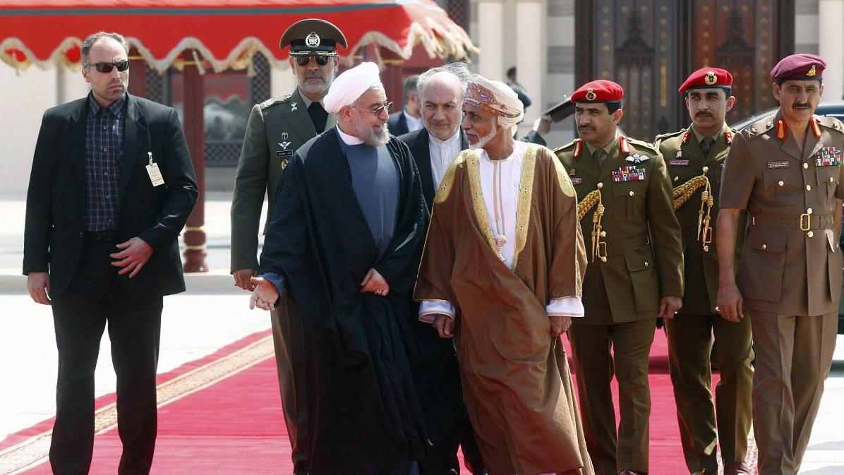 The image features a group of men walking together on a red carpet, likely in a formal or diplomatic setting. In the foreground, two men are engaged in conversation, one wearing a black suit and the other in traditional attire. They appear to be leaders or dignitaries. Behind them, there are several men in military uniforms and suits, possibly security personnel or officials, accompanying the two main figures. The background suggests an ornate architectural setting, enhancing the official nature of the gathering.