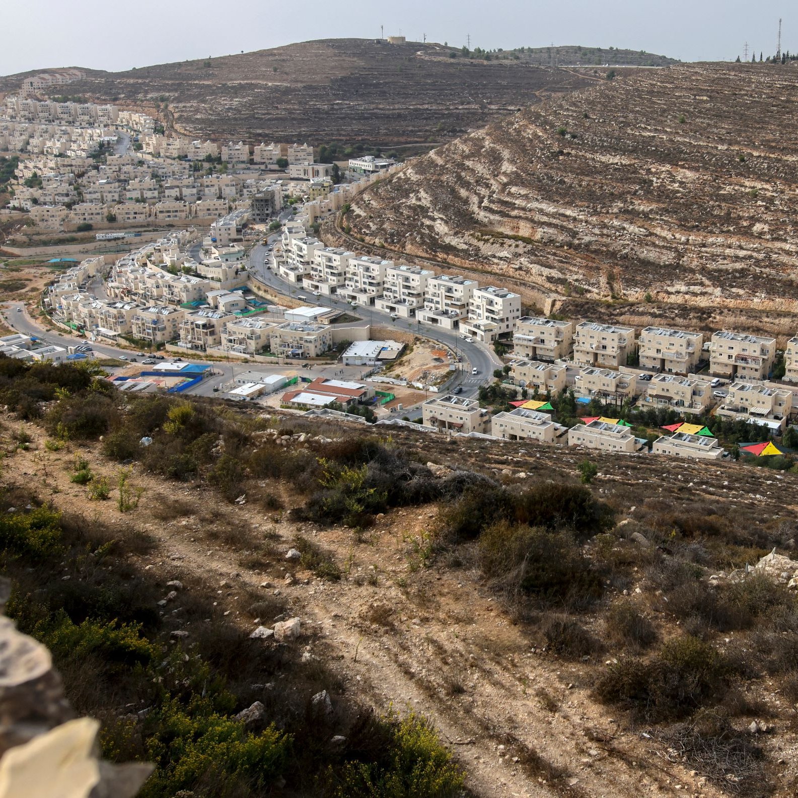 L'immagine mostra un paesaggio collinare, caratterizzato da una zona abitata con edifici bianchi disposti lungo una strada che si snoda tra le colline. In primo piano, si vede una vegetazione bassa e cespugliosa. Sullo sfondo, le colline presentano strati di roccia esposti, creando un effetto di stratificazione geologica. La luce appare naturale, suggerendo un'ora del giorno in cui il sole è alto, illuminando la scena con tonalità calde.