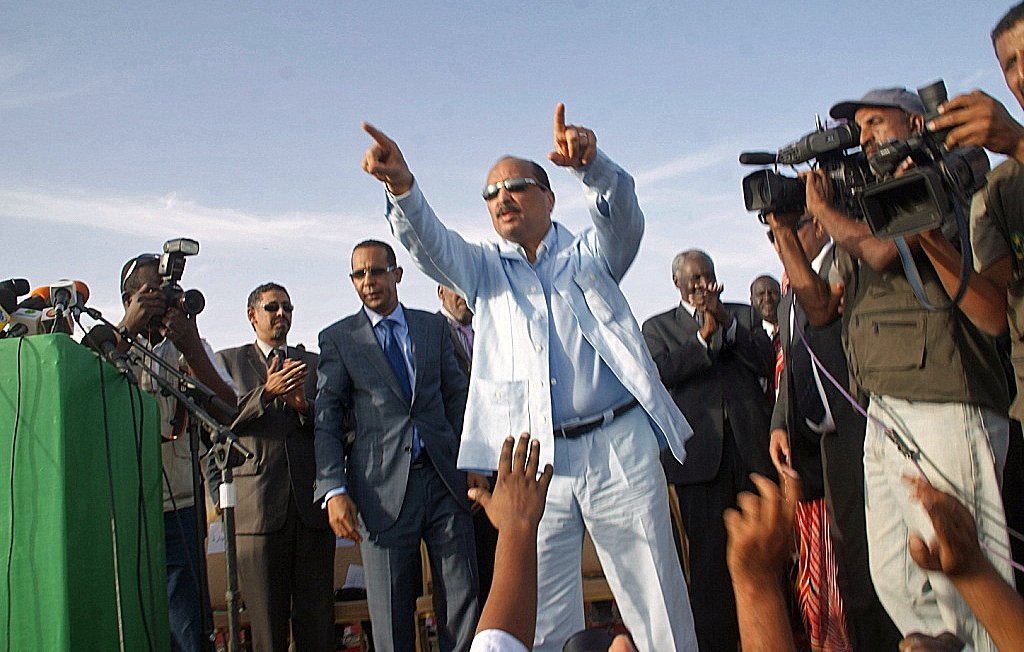 L'image montre un homme debout sur une scène, habillé en costume clair. Il semble être en train de faire un discours et de pointer vers le public. Autour de lui, plusieurs personnes l'écoutent attentivement, dont certaines sont en train de prendre des photos ou de filmer avec des caméras. Il y a une ambiance d'événement public ou politique, avec un discours énergique et engageant.