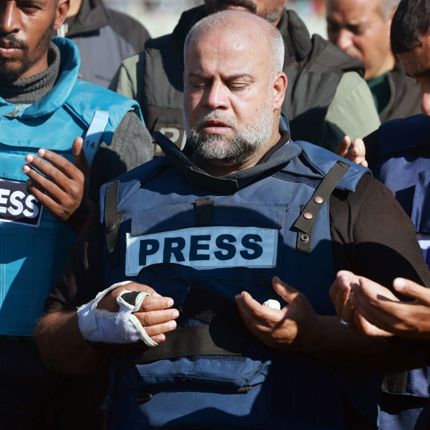 The image depicts a group of individuals, including a central figure wearing a blue vest marked "PRESS." This person appears to be in a moment of prayer or contemplation, with a serious expression, and has a bandaged hand. Surrounding him are others also dressed in similar press attire, some raising their hands in a gesture of prayer. The setting suggests a somber or reflective atmosphere, likely related to a significant event or situation involving journalists.