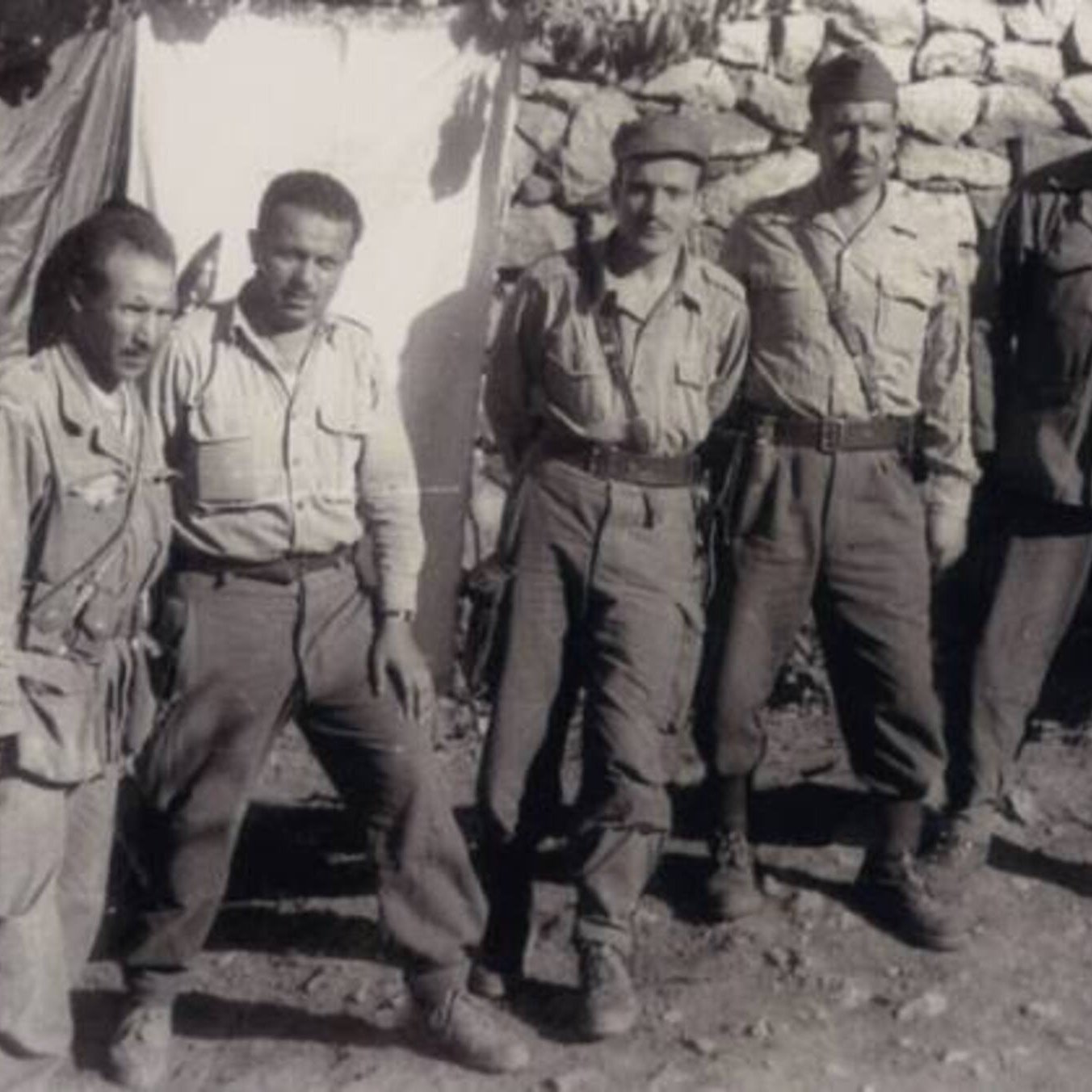 L'image présente un groupe de cinq hommes en uniforme militaire, debout ensemble devant un mur en pierres. Les hommes semblent être d'origine variée, portant des vêtements similaires, avec des pantalons et des chemises. Deux d'entre eux tiennent des armes à feu, et leur expression dégage un sentiment de camaraderie et de détermination. En arrière-plan, on aperçoit un drap suspendu, ce qui suggère qu'ils se trouvent dans un camp ou un endroit de rassemblement. L'atmosphère est sérieuse, typique des contextes militaires.