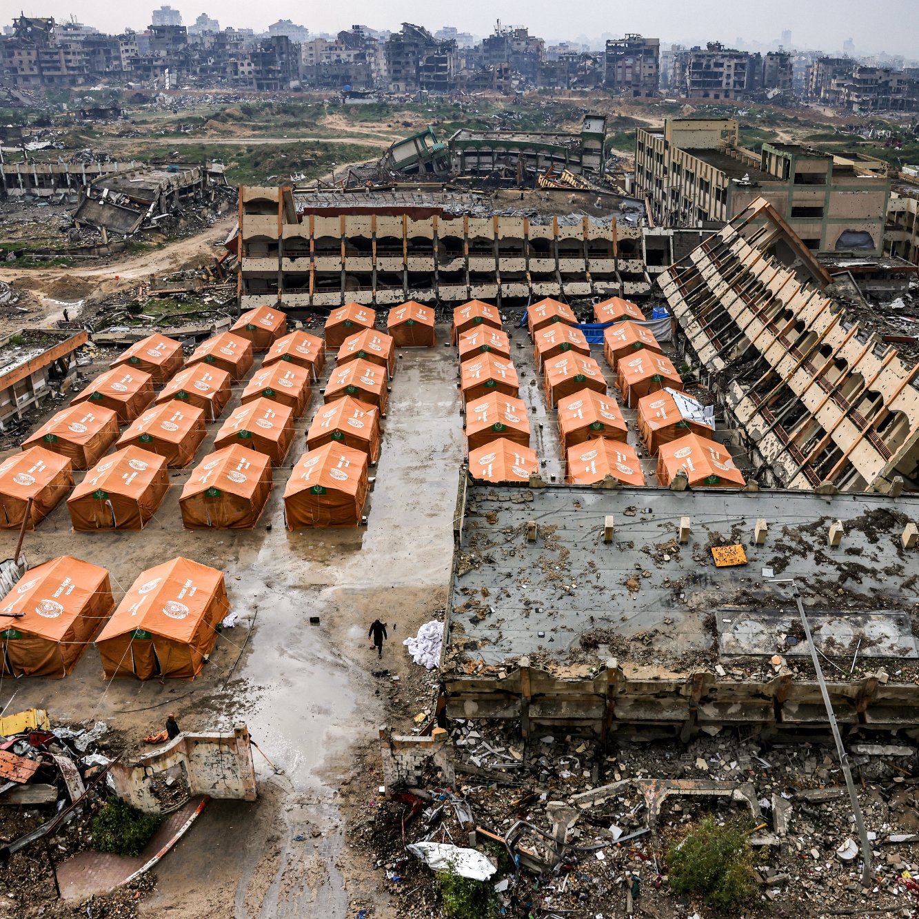 L'immagine mostra un'area devastata, con edifici in rovina sullo sfondo e tende arancioni disposte ordinatamente in primo piano. Il terreno sembra sporco e disordinato, evidenziando una situazione di emergenza o di sfollamento.