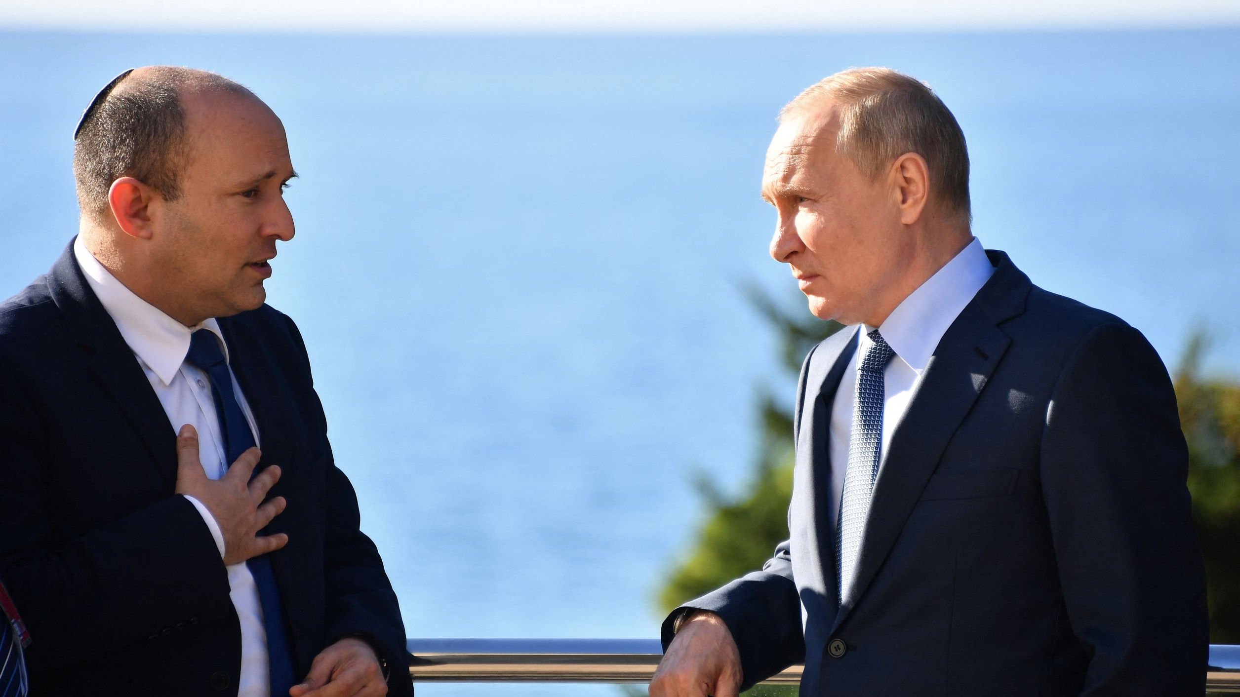 The image shows two men engaged in conversation by a seaside. One man is wearing a suit and has some form of head covering, indicating he may be of Jewish faith. The other man is also dressed in a suit and appears to be listening intently. They are both standing outdoors with a view of the water in the background. The setting suggests a formal or diplomatic meeting.