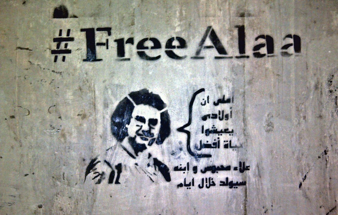 L'image présente un graffiti sur un mur. Le texte "#FreeAlaa" est inscrit en haut, accompagnant un dessin stylisé d'une personne. En dessous, il y a une citation en arabe qui semble évoquer des thèmes de liberté ou de résistance. L'ensemble donne une impression de soutien à une cause ou un individu en particulier, en soulignant la lutte pour la liberté d'expression.