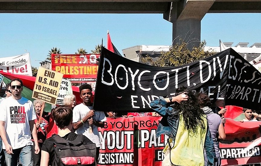 L'image montre une manifestation avec de nombreuses personnes tenant des pancartes et des bannières. Les participants semblent exprimer des opinions politiques concernant Israël et la Palestine. On peut lire des slogans comme "Boycott, Désinvestissez, Sanctionnez" et "Finissez avec Israël". L'ambiance paraît dynamique, avec des gens engagés et un sentiment d'unité pour une cause. La scène se déroule sous un viaduc, ajoutant au cadre urbain de l'événement.
