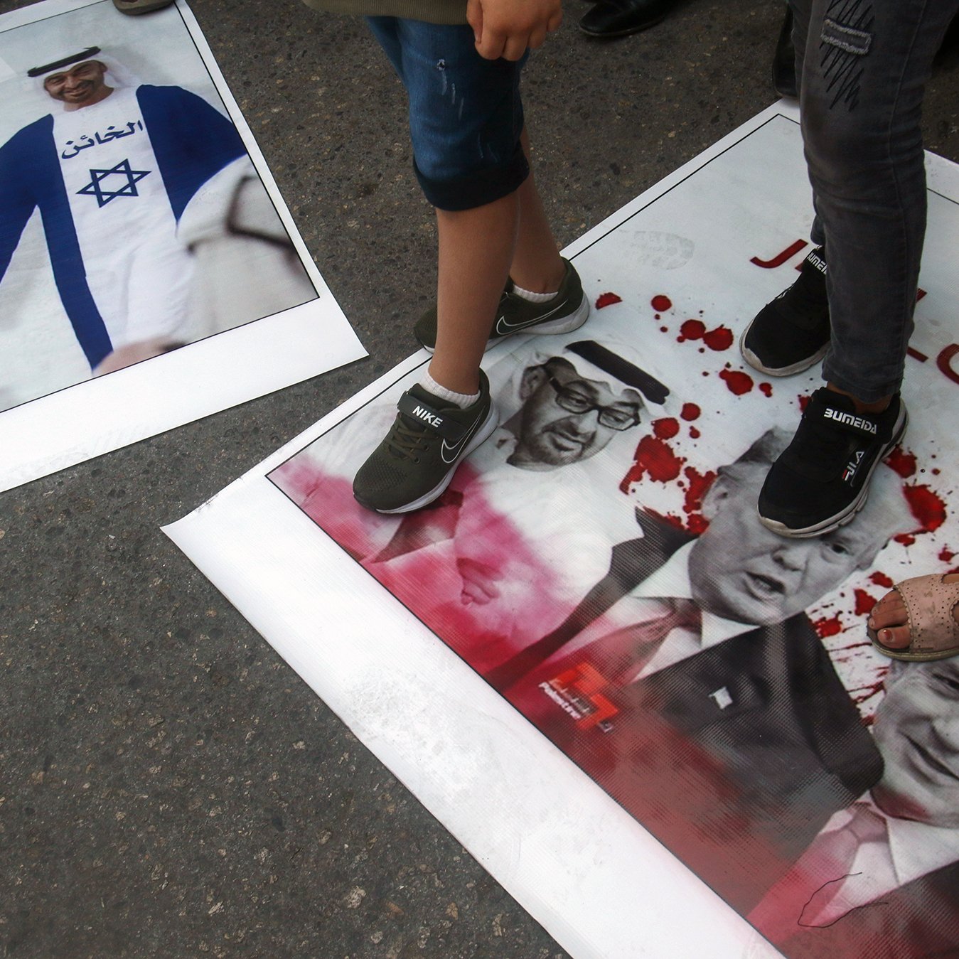L'image montre des affiches au sol sur lesquelles sont piétinées des images de personnalités politiques, notamment Benjamin Netanyahu, Donald Trump et d'autres figures. Les affiches semblent être liées à une manifestation ou une protestation, avec des couleurs vives et des motifs qui attirent l'attention. Les personnes présentes dans l'image sont en train de marcher dessus, suggérant une attitude de rejet ou de mécontentement envers ces figures. Les tonnerres de couleurs et le contenu des affiches pourraient refléter des sentiments politiques forts.