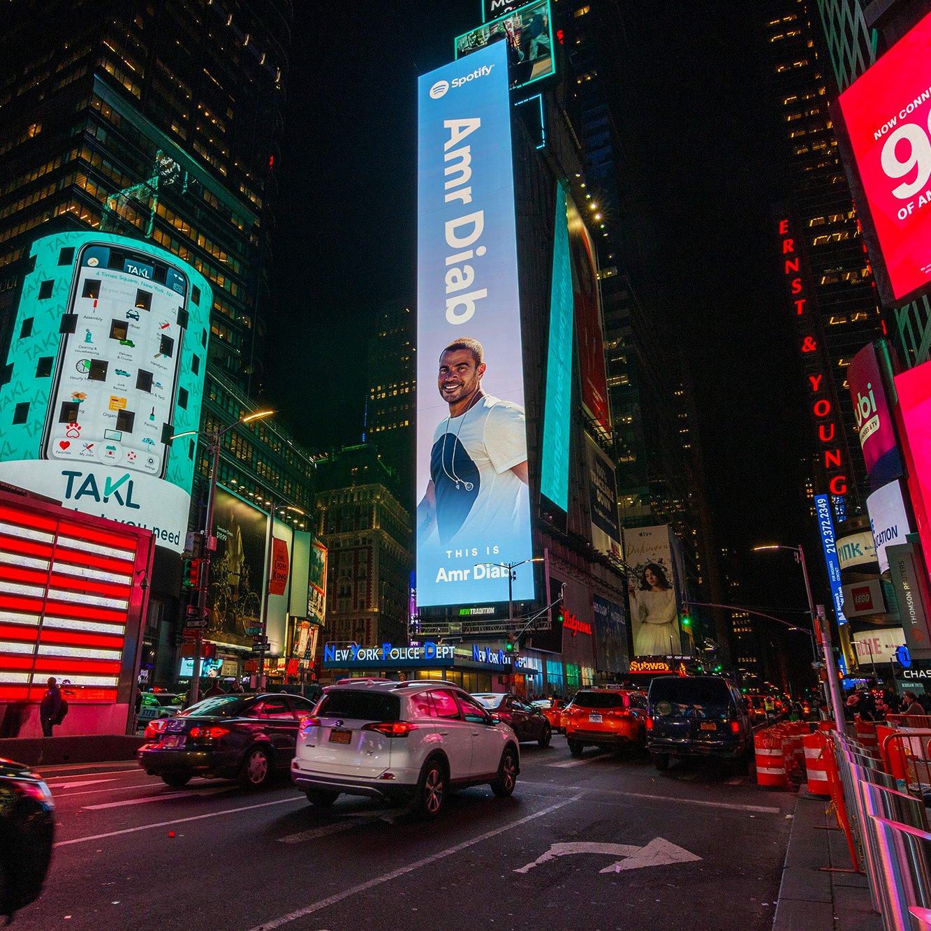The image depicts a vibrant scene in Times Square, New York City, at night. The area is illuminated by numerous bright advertisements and digital billboards. Prominently featured is a large advertisement for Amr Diab, an acclaimed musician, which invites viewers to listen to his music. There is an American flag displayed on one of the buildings, and several vehicles are seen navigating the busy street. The atmosphere is lively, characteristic of this iconic urban location.