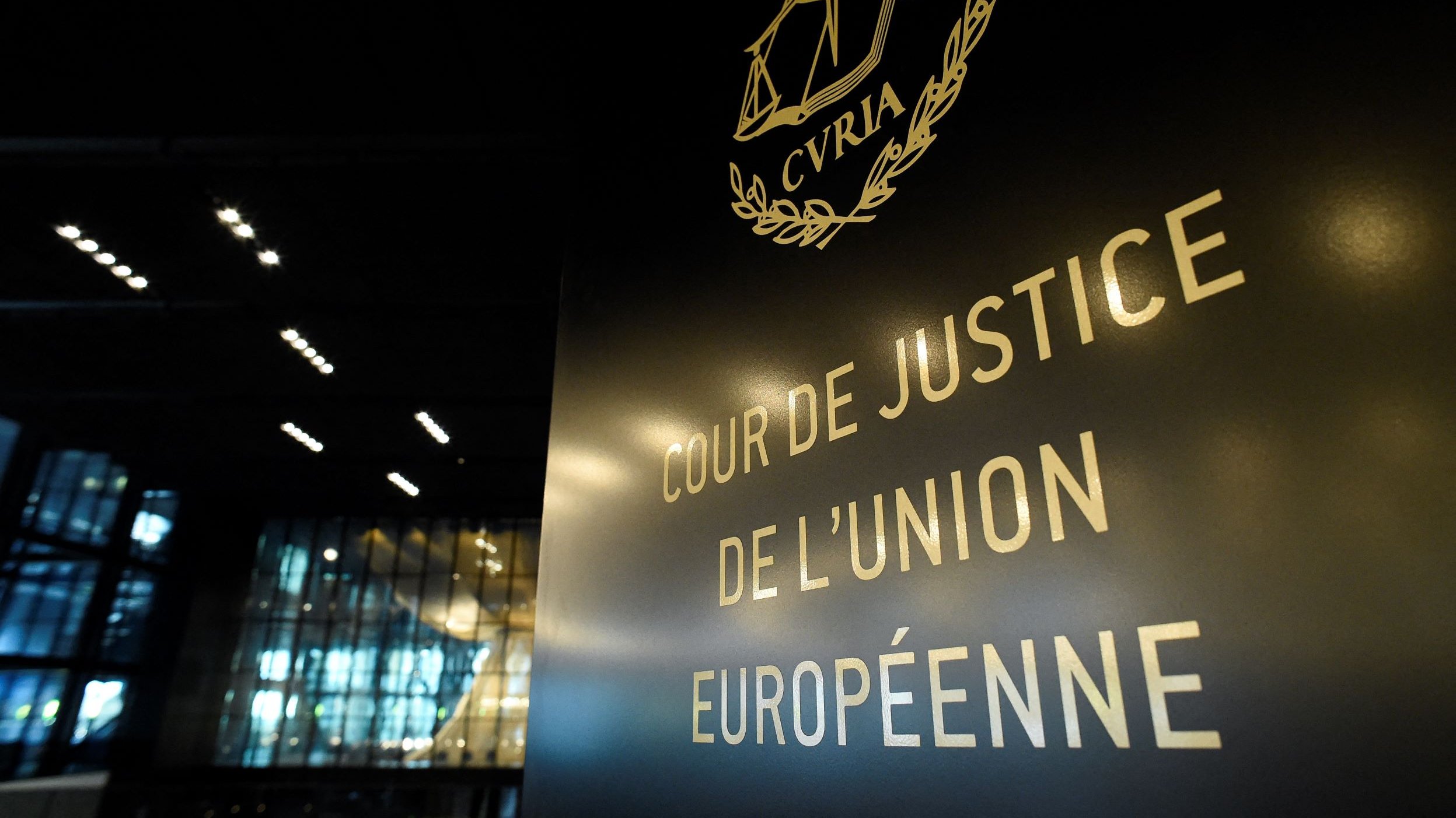 L'image montre un panneau indiquant "COUR DE JUSTICE DE L'UNION EUROPÉENNE". Le panneau est stylisé avec des éléments graphiques, notamment une représentation de la balance de la justice. En arrière-plan, on peut voir une architecture moderne et intérieure, probablement d'un bâtiment officiel. L'éclairage est subtil, mettant en valeur le texte et le design du panneau.