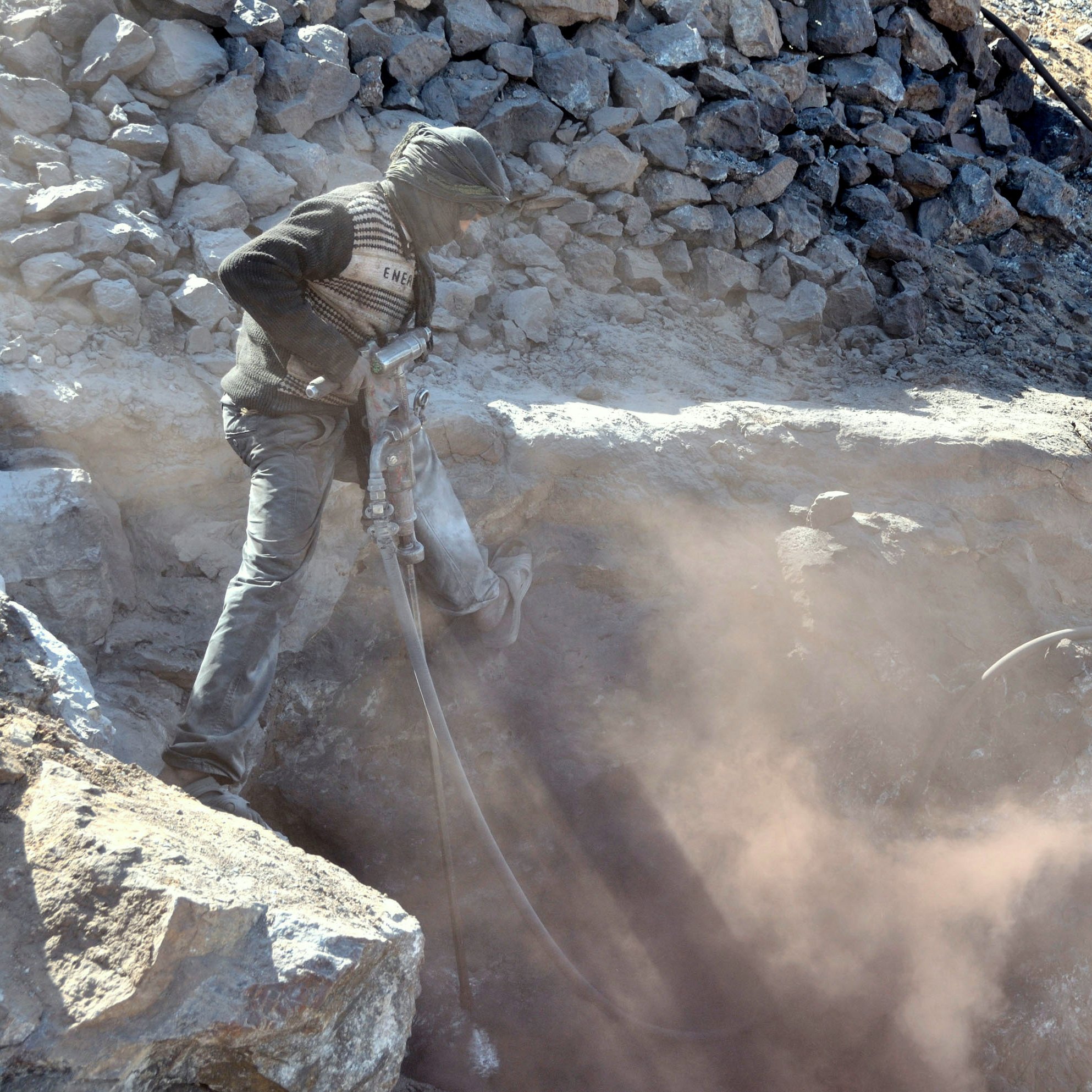 The image depicts a worker operating a pneumatic drill in a rocky area. The person is wearing protective clothing, including a mask or scarf over their face to shield against dust. Surrounding the worker are loose rocks and debris, and a cloud of dust is being kicked up by the drilling activity, creating a hazy atmosphere. The setting appears to be a mining or construction site.