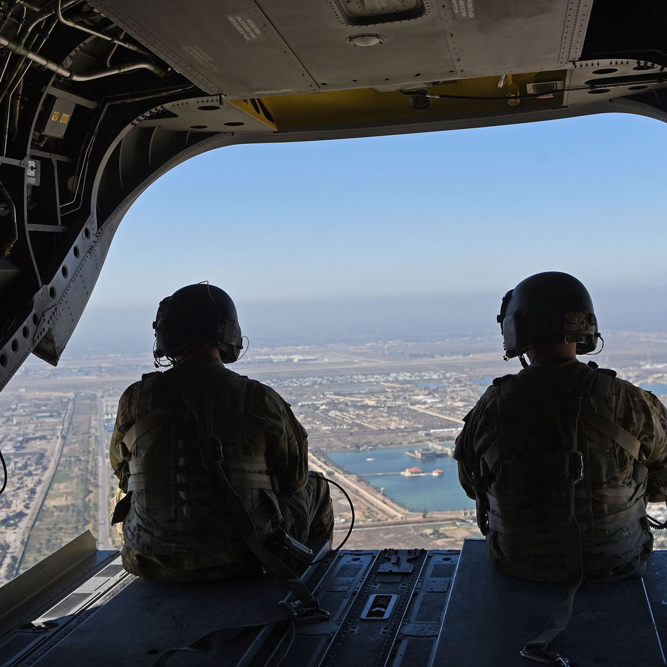 L'image montre deux membres d'équipage militaires assis à l'arrière d'un hélicoptère, regardant à l'extérieur. Ils portent des uniformes de camouflage et des casques. À travers l'ouverture de l'appareil, on peut apercevoir un paysage urbain et un plan d'eau en contrebas, sous un ciel clair. La vue offre un aperçu de la ville avec des routes et des bâtiments visibles.