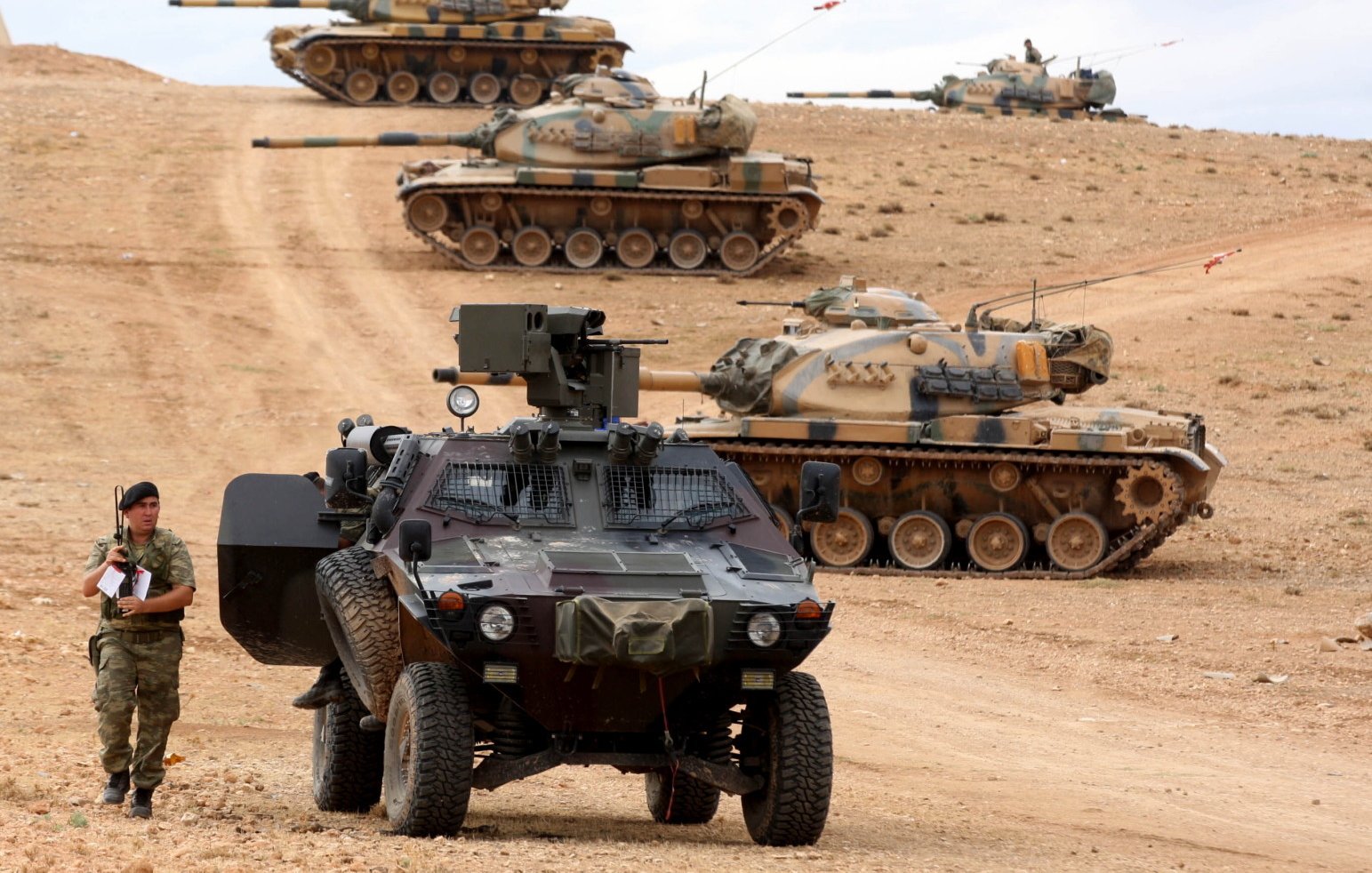 L'image montre un paysage militaire avec plusieurs chars d'assaut en arrière-plan, alignés sur une colline. Au premier plan, un véhicule blindé est garé sur un chemin poussiéreux, tandis qu'un soldat semble faire une inspection ou prendre des notes à côté du véhicule. Le terrain est désertique, et le ciel est partiellement nuageux, créant une atmosphère tendue et militaire. Les chars sont camouflés avec des motifs de peinture qui se fondent dans le paysage environnant.