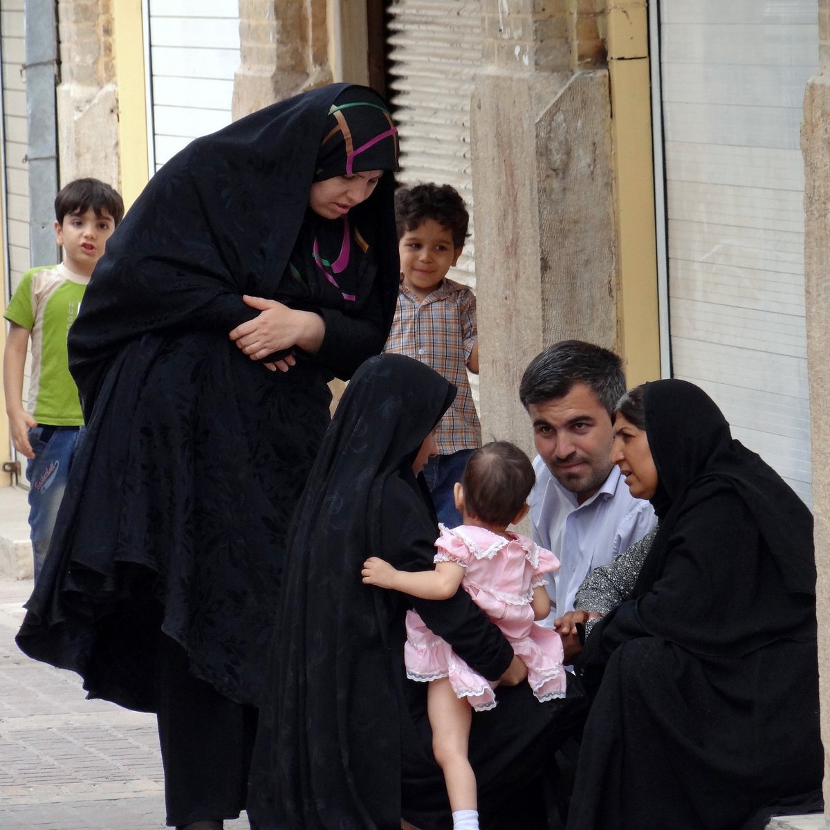 L'image montre un groupe de personnes dans une rue. Deux femmes, vêtues de vêtements noirs, sont assises sur le sol et discutent. L'une d'elles tient un bébé, qui porte une légère robe rose. À proximité, un homme regarde les femmes et semble participer à la conversation. Un jeune garçon, en arrière-plan, observe la scène. L'environnement apparaît urbain, avec des bâtiments en briques visibles. L'atmosphère semble conviviale et familiale.