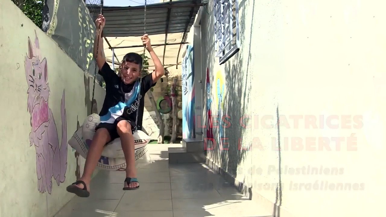 L'image montre un jeune garçon qui se balançe sur une balançoire improvisée. Il semble joyeux et se trouve dans un espace qui semble être un couloir ou une cour. Les murs autour de lui sont décorés avec des peintures murales, dont une représente un chat. Le cadre donne une impression de vie quotidienne dans un endroit où l'art et le jeu se côtoient. On voit également un message en arrière-plan, lié à des thèmes de liberté.