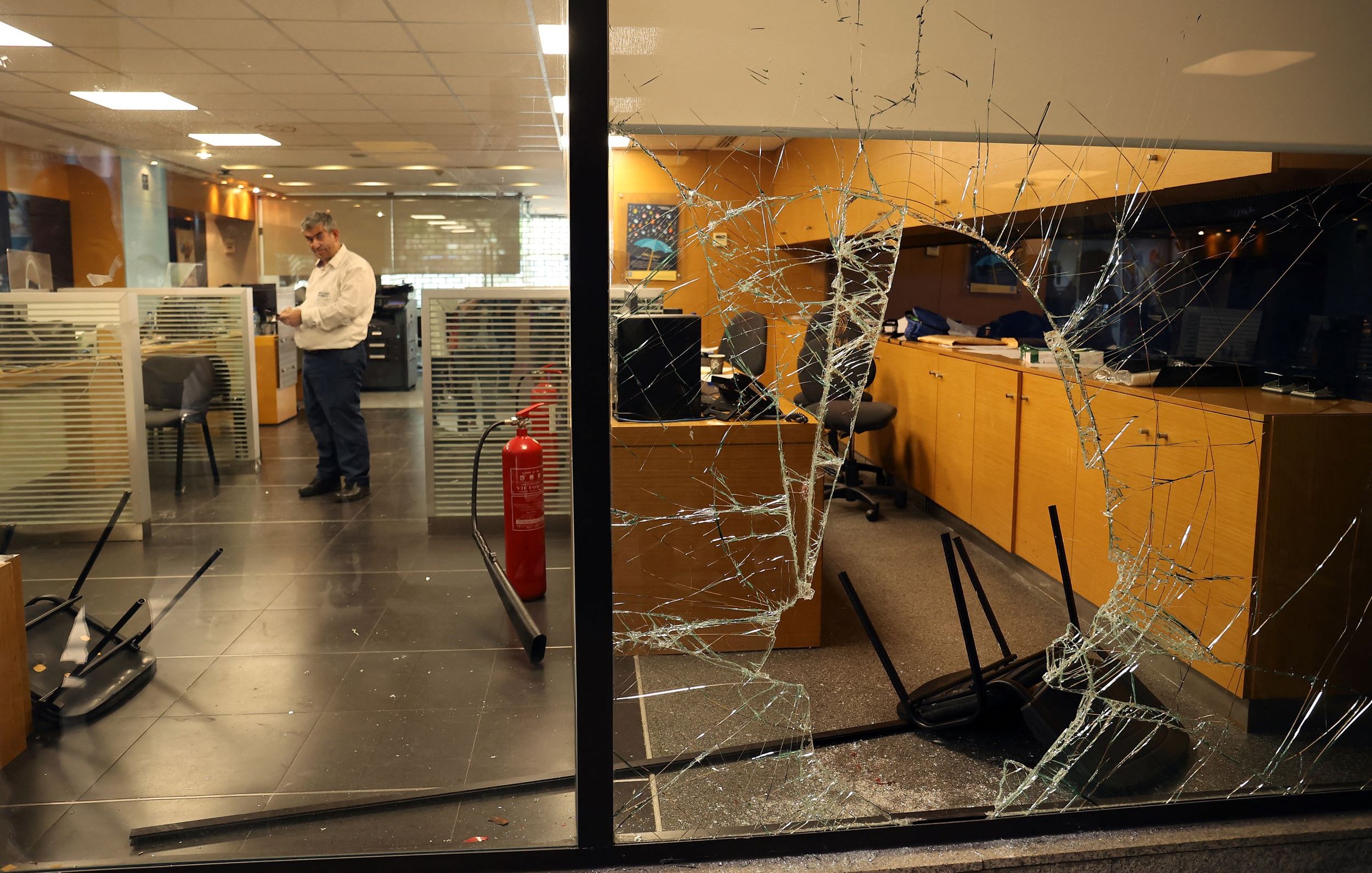 L'image montre un bureau avec des dégâts visibles. On peut voir une vitre brisée à l'avant, avec des fissures qui s'étendent sur toute sa surface. À l'intérieur, un homme est debout, semblant examiner la situation, tandis que des chaises sont renversées sur le sol. L'ambiance générale suggère qu'il y a eu un incident perturbateur dans cet espace de travail.