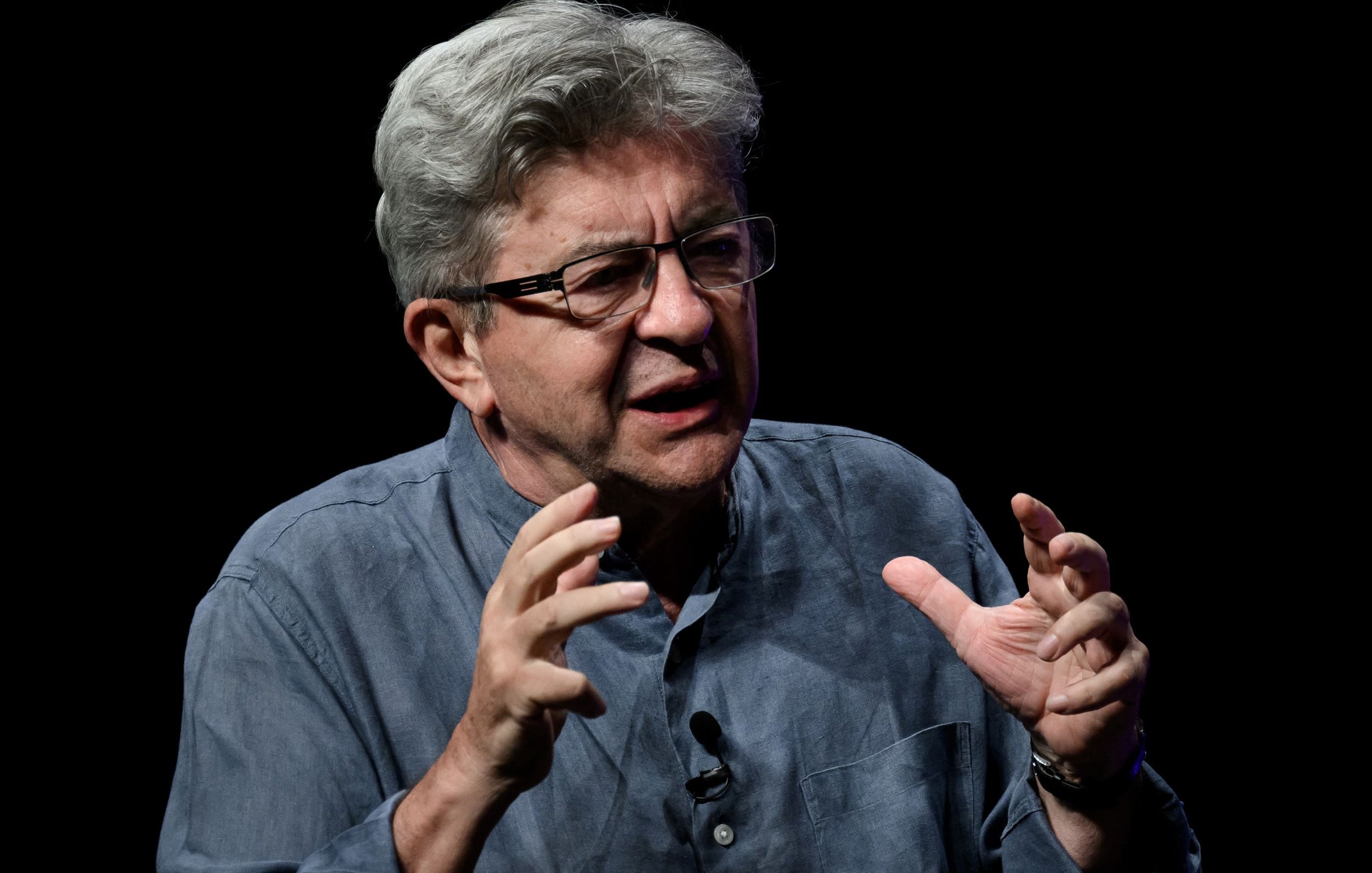 این تصویر یک مرد میانسال را نشان می‌دهد که به نظر می‌رسد در حال صحبت کردن یا بحث کردن است. او عینک به چشم دارد و موهای خاکستری و حالت نامنظمی دارد. لباس او به رنگ آبی تیره یا خاکستری است و به نظر می‌رسد که در یک محیط تاریک یا با پس‌زمینه‌ای سیاه قرار دارد. او دست‌هایش را در حالت ابراز احساسات یا تأکید بر نقاط صحبت خود حرکت می‌دهد.