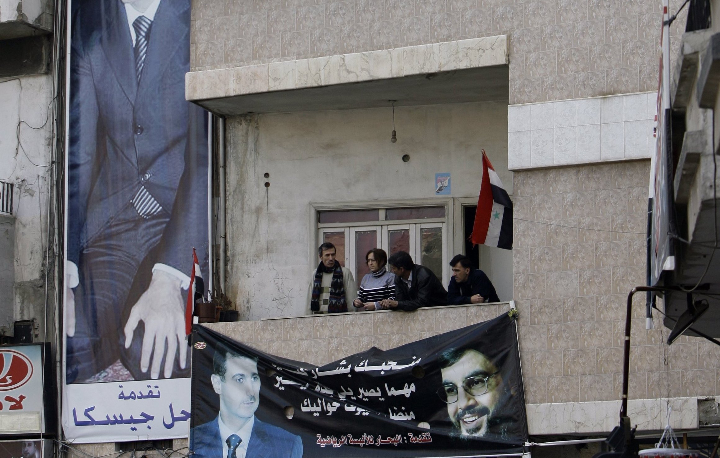 L'image montre un bâtiment avec une grande affiche représentant un homme en costume, probablement une figure politique. On peut voir plusieurs personnes sur un balcon au premier étage, observant l'extérieur. En dessous, il y a une bannière affichant un message, et des drapeaux sont visibles, ce qui suggère un contexte politique ou de soutien à une cause particulière. Les murs du bâtiment sont décorés de manière simple, et l'ambiance semble être formelle.