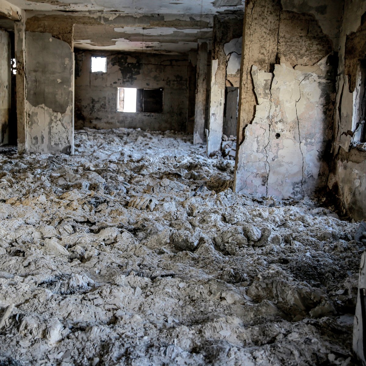 تظهر الصورة غرفة داخل مبنى تضرر بشكل كبير، حيث الجدران والسقف متهدمان، والأرضية مغطاة بطبقة من المواد البيضاء، مما يوحي بتدهور شديد في حالة المبنى. يدخل الضوء من بعض النوافذ المكسورة، مما يضيف شعورًا بالخراب والمهجورية إلى المكان.