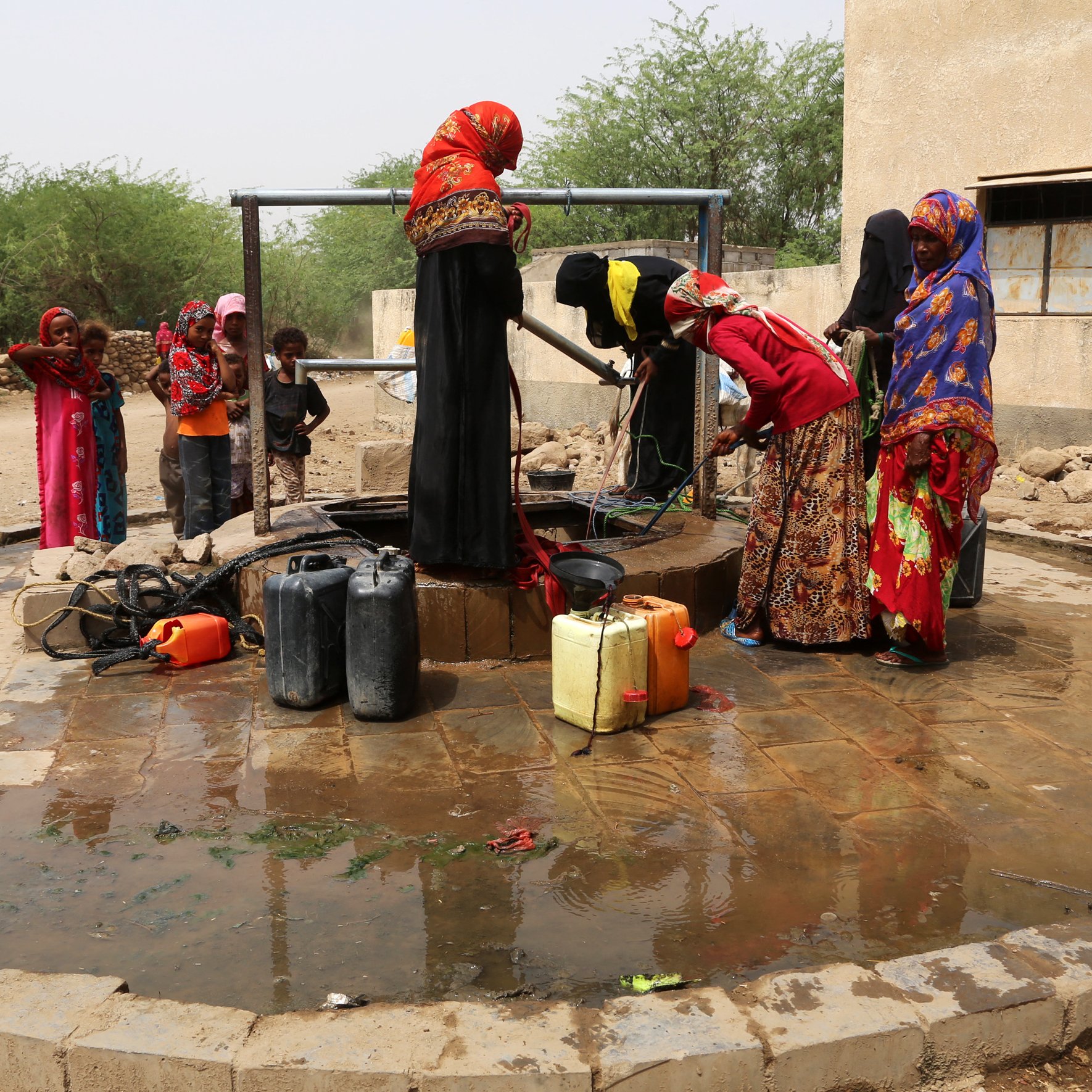 L'image montre un groupe de femmes près d'un puits. Elles sont vêtues de robes colorées et semblent être en train de remplir des conteneurs d'eau. Des enfants jouent autour d'elles. Le sol est humide, et le cadre semble rural, avec des buissons et une structure en arrière-plan. L'atmosphère semble active et communautaire, illustrant l'importance de l'accès à l'eau dans la vie quotidienne.
