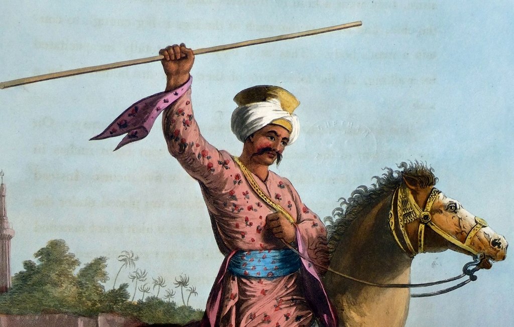 L'image représente un cavalier en tenue traditionnelle. Il porte un turban et des vêtements colorés ornés de motifs, avec une ceinture large. Le cavalier est en train de tenir un long bâton levé, semblant exprimer force et détermination. Il est monté sur un cheval puissant, qui avance au galop. En arrière-plan, on peut apercevoir d'autres cavaliers, ce qui suggère une scène dynamique. L'environnement semble indiquer un paysage désertique ou aride, renforçant le caractère historique de l'image.