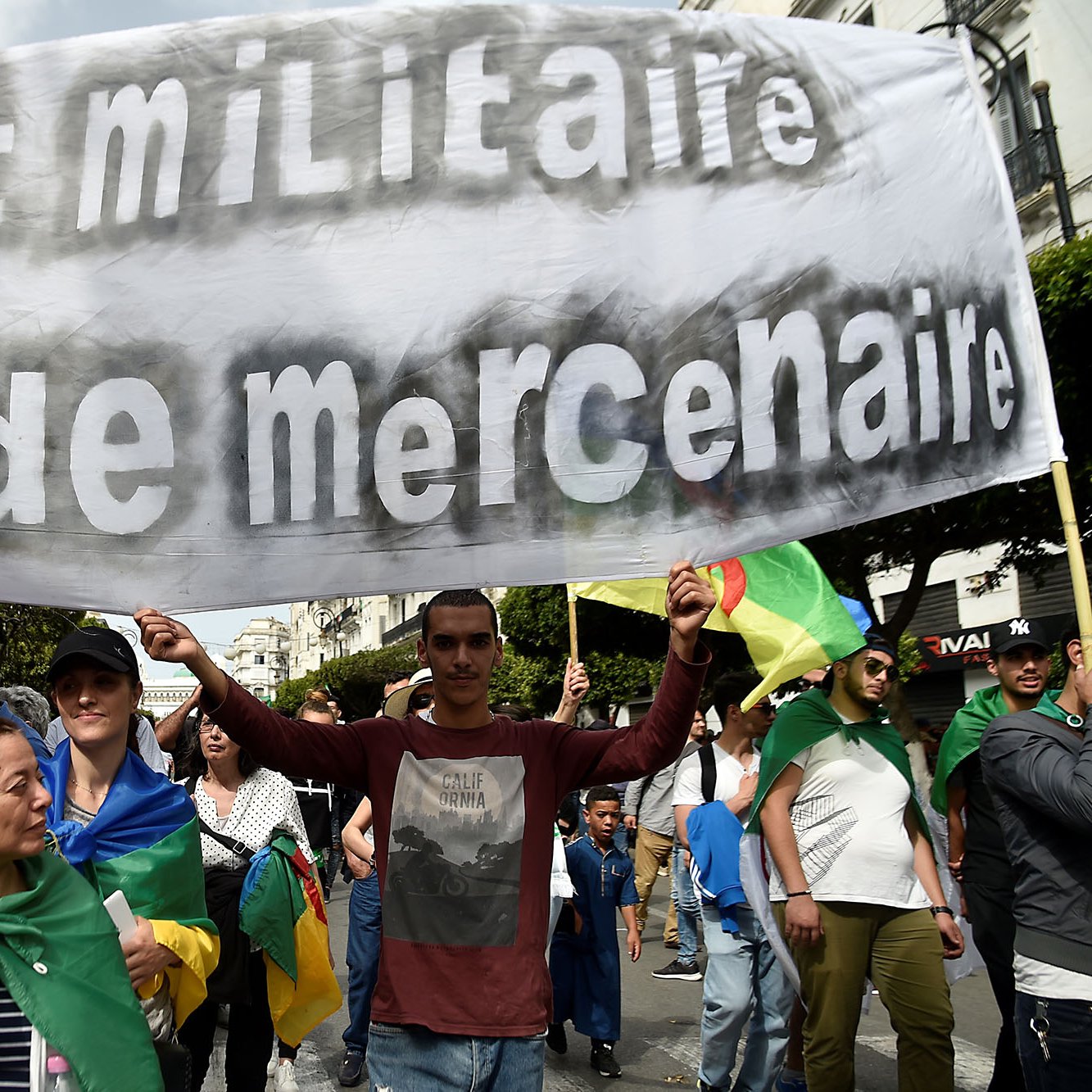 تصویر مربوط به یک تظاهرات است که در آن جمعیتی از مردم در حال حرکت هستند و یک بنر بزرگ را در دست دارند. بنر نوشته‌ای به زبان فرانسوی دارد که به معنای "نه دولت نظامی، نه باند مزدور" است. افراد حاضر در تظاهرات به نظر می‌رسد از اقشار مختلف جامعه هستند و در پس‌زمینه، فضای شهری قابل مشاهده است. برخی از تظاهرکنندگان پرچم‌هایی به دوش دارند و جو کلی بر آزادی‌خواهانه و اعتراضی تمرکز دارد.