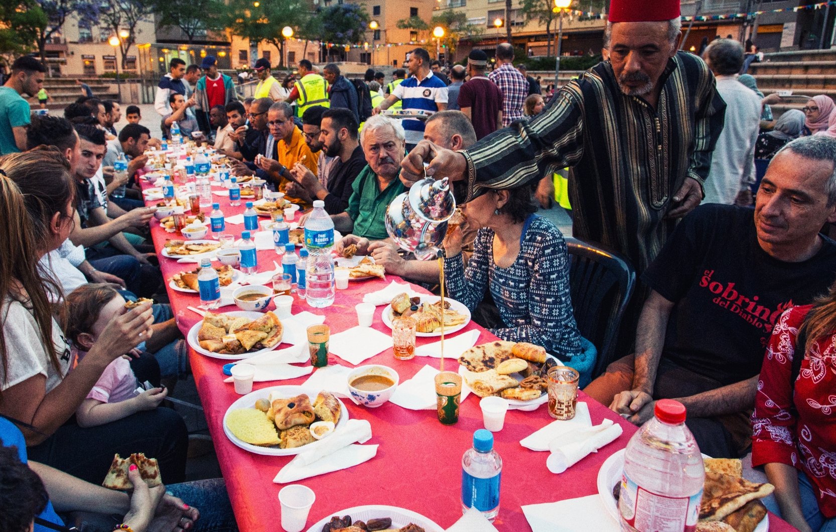 L'image montre une grande table dressée en plein air, où des personnes diverses sont réunies autour d'un repas convivial. Les convives, de différents âges, sont assis sur des chaises en plastique et profitent de divers plats, avec des assiettes remplies de nourriture. On peut voir des bouteilles d'eau sur la table et des verres. L'ambiance semble festive et chaleureuse, marquée par des sourires et des échanges entre les participants. En arrière-plan, on aperçoit d'autres personnes et des éléments de l'environnement urbain, indiquant une atmosphère de communauté.