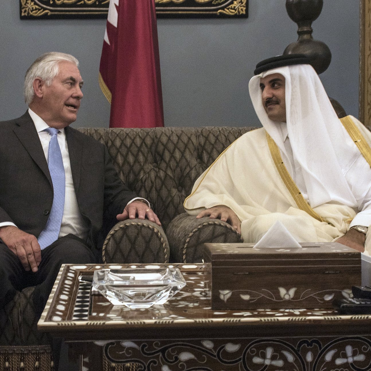 L'image montre deux hommes assis dans un cadre élégant. L'un d'eux porte un costume noir avec une cravate claire, tandis que l'autre est habillé d'une tunique traditionnelle blanche et d'un manteau beige. Ils semblent engagés dans une conversation conviviale, entourés de décorations raffinées, y compris des coussins rouges et des ornements sur la table. L'atmosphère est de camaraderie et de diplomatie.