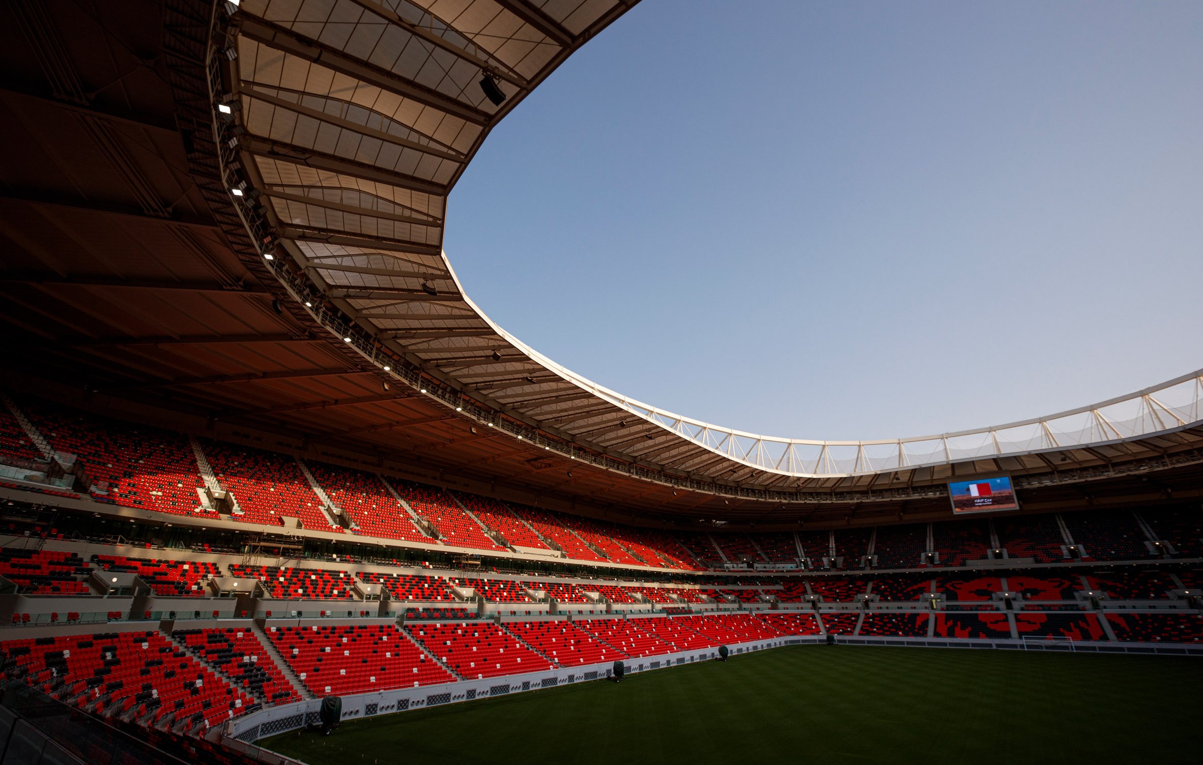 L'image montre un stade moderne avec des sièges rouges et une large toiture. La pelouse est bien entretenue, et on peut apercevoir un écran géant à une extrémité. Le ciel est dégagé, indiquant une belle journée. L'intérieur du stade semble prêt à accueillir un événement, avec une atmosphère calme et anticipative.