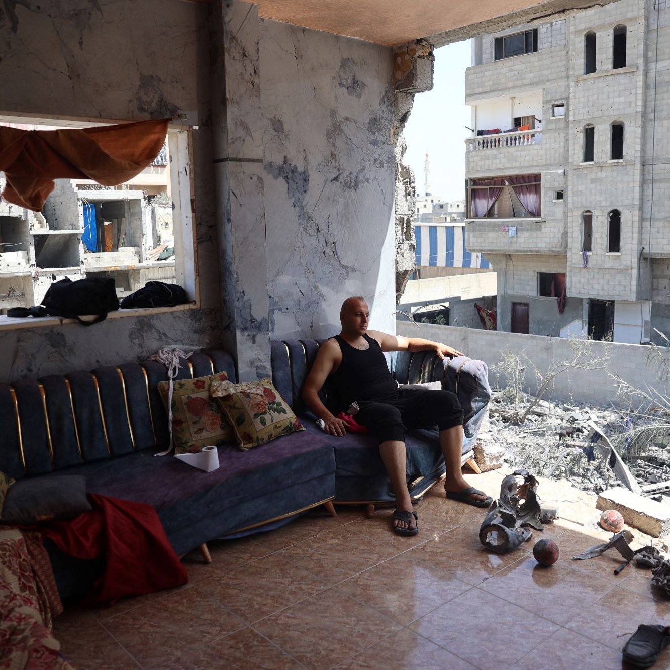 L'image montre un homme assis sur un canapé dans une pièce endommagée. Il semble se reposer, vêtu d'un débardeur noir et d'un pantalon court et noir. La pièce est en mauvais état, avec des murs endommagés et des débris au sol. À l'extérieur de la pièce, on aperçoit des bâtiments gravement endommagés, suggérant qu'il s'agit d'une zone touchée par un conflit ou une catastrophe. Le mobilier à l'intérieur, comme le canapé et certains coussins, semble également en désordre. L'atmosphère générale est celle de la dévastation et de la récupération après un événement destructeur.