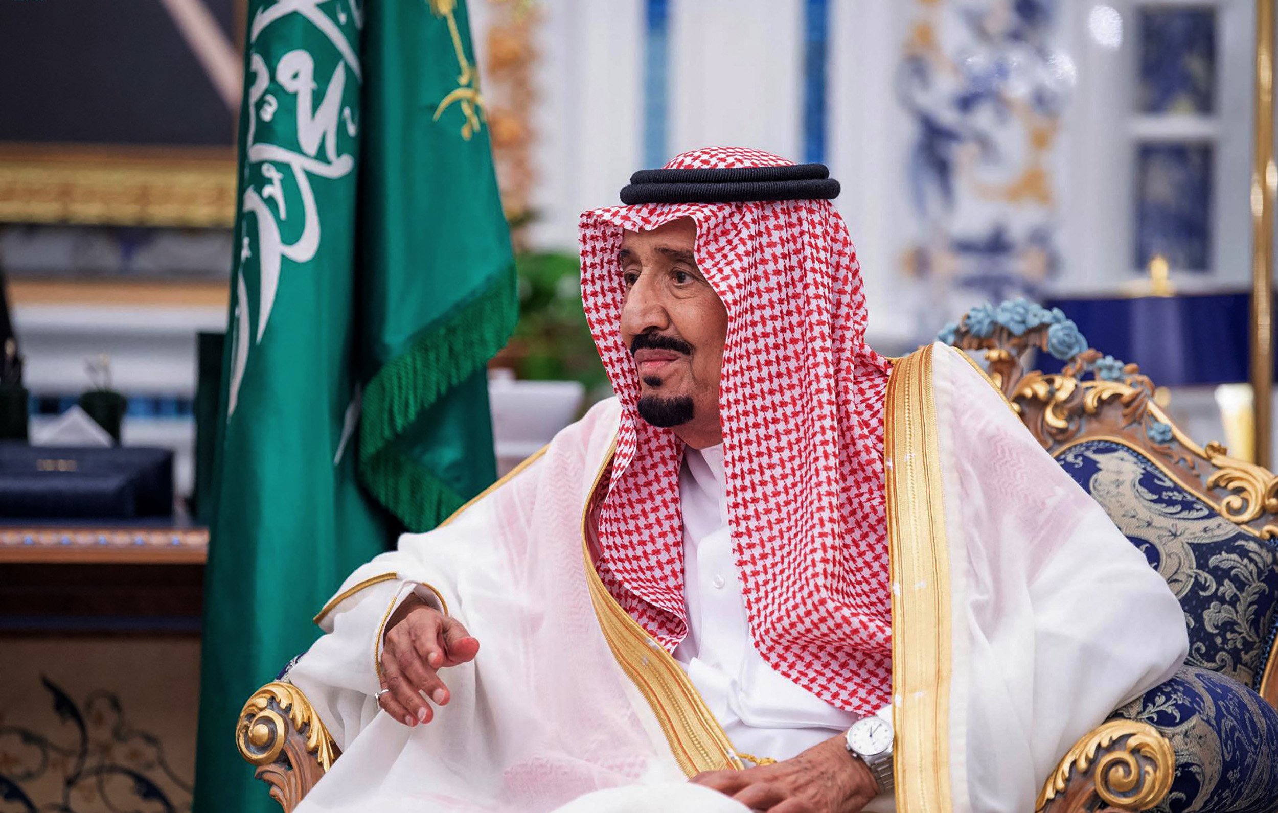 تظهر في الصورة شخصية ترتدي ملابس تقليدية سعودية، تتضمن الإزار والعباءة، كما أن هناك شالًا على رأسه. يجلس على كرسي فخم في بيئة ذات تفاصيل تراثية. خلفه توجد علم المملكة العربية السعودية، مما يدل على السياق الرسمي والمناسبات المهمة. تعكس تعابير وجهه تركيزًا واهتمامًا، كما يظهر الاهتمام بالتصميم والزخارف المحيطة به.