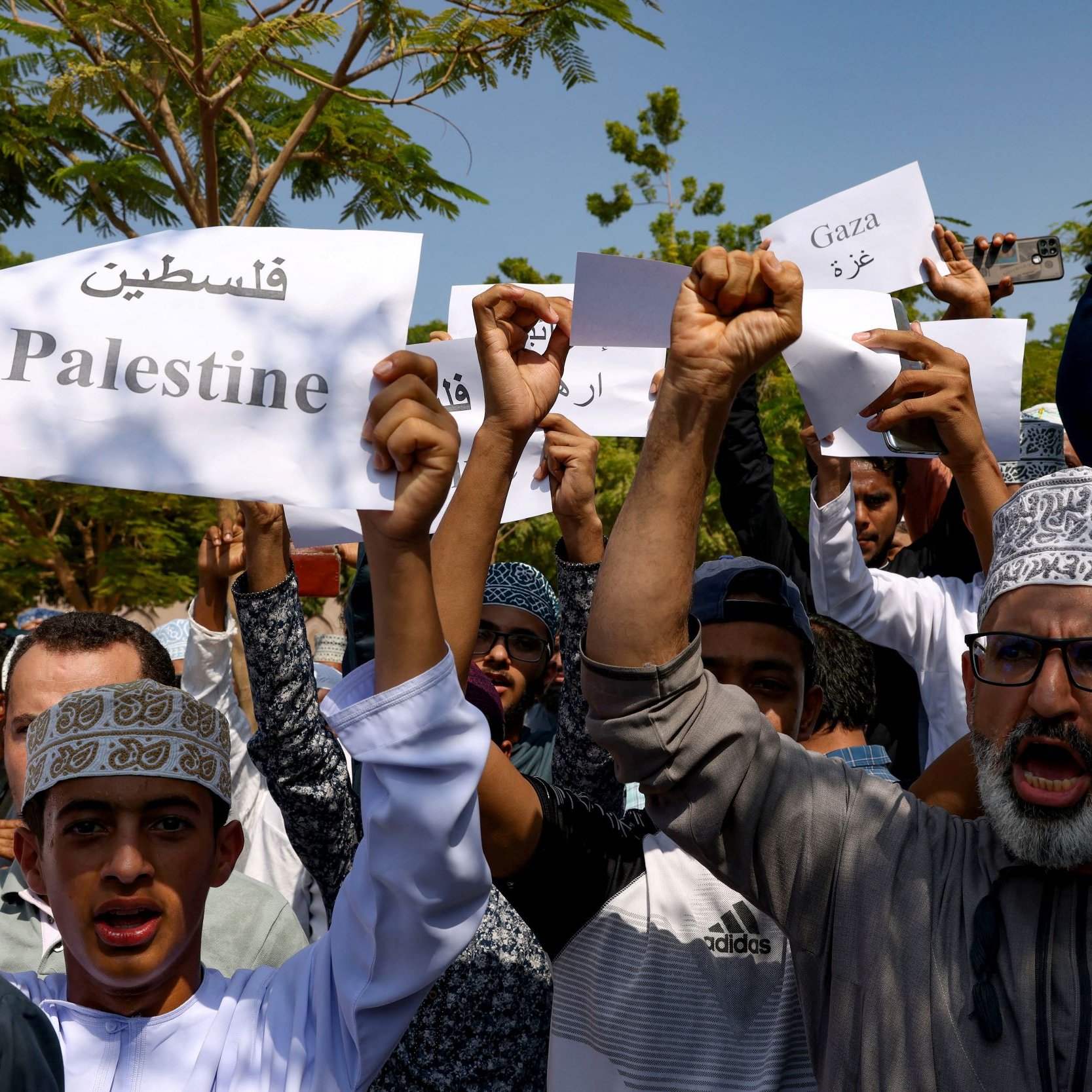L'image montre un groupe de personnes tenant des pancartes en soutien à la Palestine et à Gaza. Les manifestants semblent rassembler pour exprimer leur solidarité, avec des expressions déterminées et des chants. Les pancartes affichent des mots en arabe ainsi qu'en anglais, reflétant un message de soutien à la cause palestinienne. L'environnement semble être en plein air, probablement lors d'une manifestation ou d'un rassemblement.