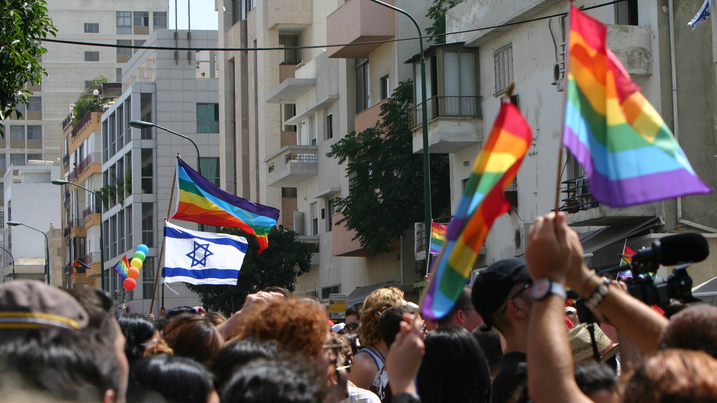 L'image montre une scène vibrante de célébration lors d'un défilé de fierté. On y voit une foule de personnes brandissant des drapeaux arc-en-ciel, symboles de la communauté LGBTQ+. Parmi eux, une grande bannière représentant le drapeau d'Israël flotte également. En arrière-plan, on distingue des bâtiments urbains, et plusieurs personnes semblent s'amuser et célébrer l'événement, créant une ambiance festive et joyeuse.
