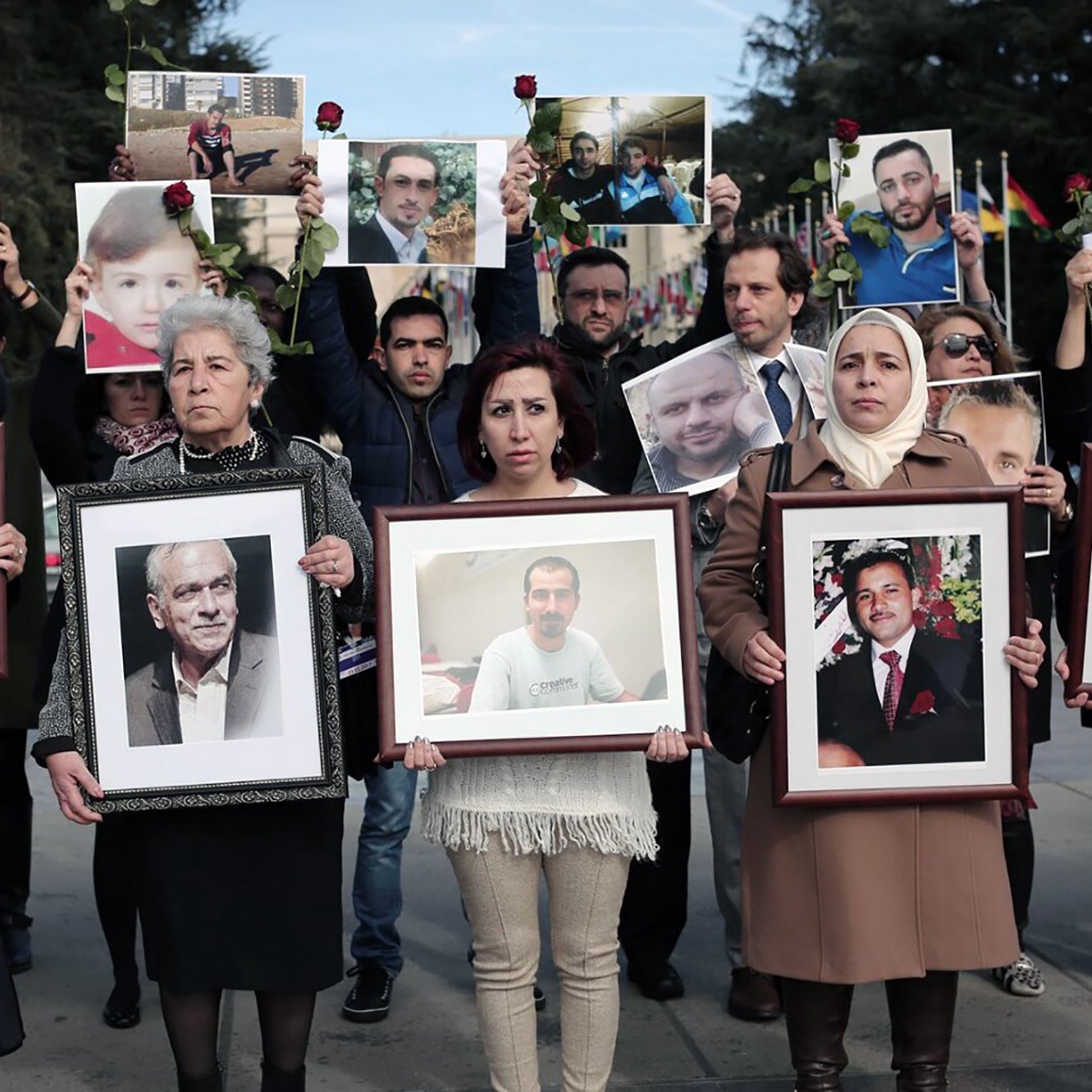 L'image montre un groupe de personnes participant à une manifestation ou un hommage. Elles tiennent des portraits de différents individus, certains avec des roses. Les expressions des participants semblent sérieuses et engagées, suggérant qu'ils rendent hommage à des personnes disparues ou aux victimes d'un conflit. Le décor en arrière-plan semble être en plein air, et plusieurs drapeaux sont visibles. Les vêtements des manifestants varient, avec quelques femmes portant des foulards.