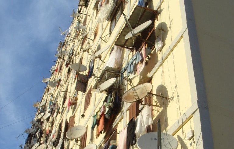 L'image montre un bâtiment d'habitation avec de nombreux balcons. Les murs de l'immeuble sont couverts de vêtements suspendus à sécher, créant un décor coloré et animé. De plus, plusieurs antennes parabolique sont fixées sur les façades, indiquant que les habitants ont accès à la télévision satellite. Le ciel est clair, ajoutant une lumière naturelle à la scène. L'ensemble donne une impression de vie communautaire et de simplicité dans un cadre urbain.