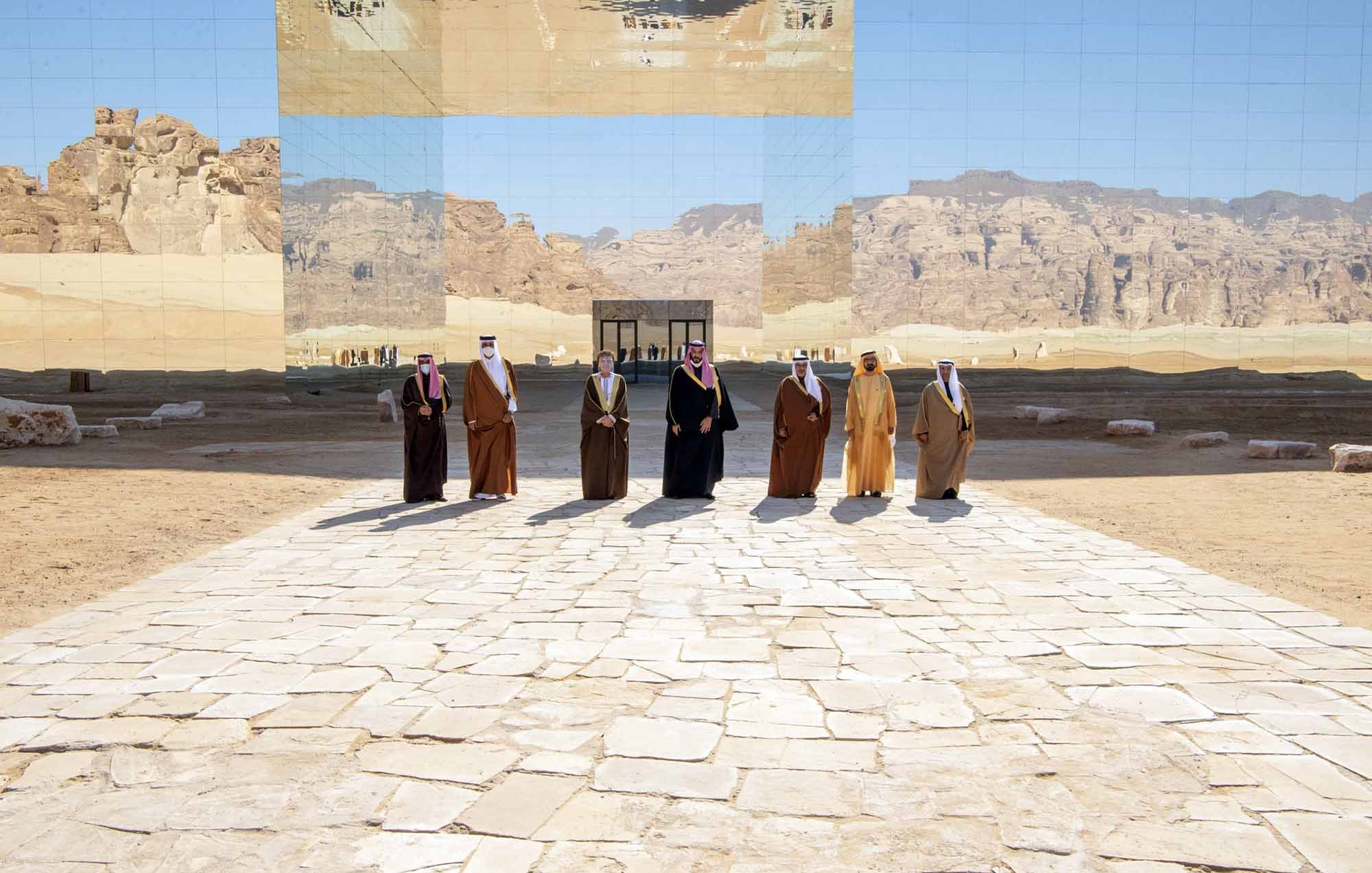 تظهر في الصورة مجموعة من الأشخاص يرتدون الزي التقليدي، ويقفوا على طريق مرصوف بالحجارة في منطقة صحراوية. خلفهم توجد جدران عاكسة تعكس المنظر الجبلي المحيط، مما يعطي شعوراً بالتواصل بين البشر والطبيعة. السماء زرقاء وصافية، مما يضيف جمالاً على المشهد.