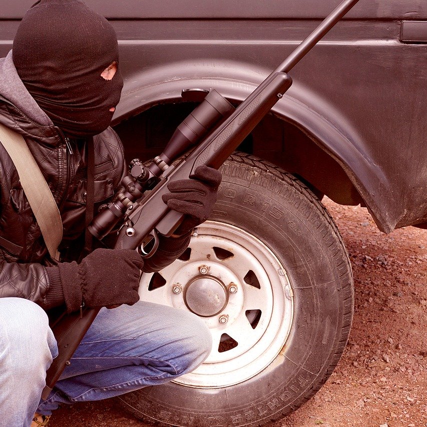 L'image représente une personne en position accroupie, portant un masque noir qui couvre son visage. Elle est habillée d'une veste en cuir et de gants noirs. Dans ses mains, elle tient un fusil équipé d'un accessoire de visée. En arrière-plan, on peut voir une camionnette avec une roue visible, suggérant un environnement extérieur. L'ensemble dégage une atmosphère tendue et dramatique.