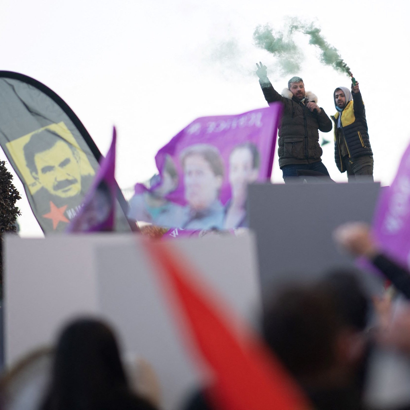 L'image montre une scène de manifestation. Des personnes brandissent des drapeaux et des bannières colorées, principalement en violet et rouge. Au premier plan, on aperçoit deux personnes sur une estrade, l'un d'eux tenant un mégaphone et levant les bras, tandis que l'autre semble encourager la foule. De la fumée verte s'échappe, ajoutant une atmosphère dynamique à l'événement. En arrière-plan, on distingue des portraits, probablement de figures politiques ou symboliques. L'ensemble dégage une ambiance de mobilisation et de passion.