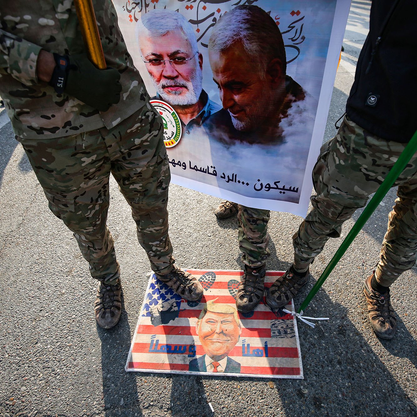 L'image montre des soldats en uniforme militaire debout sur une affiche ou un symbole qui semble représenter des sentiments anti-américains. Ils tiennent des drapeaux ou des bannières, et il y a une image de personnes, probablement des figures politiques ou militaires, sur l'affiche. Le sol est décoré d'une illustration de style artistique, mélangé avec des éléments du drapeau américain, ce qui suggère une forte opinion politique et un contexte de protestation ou de commémoration.