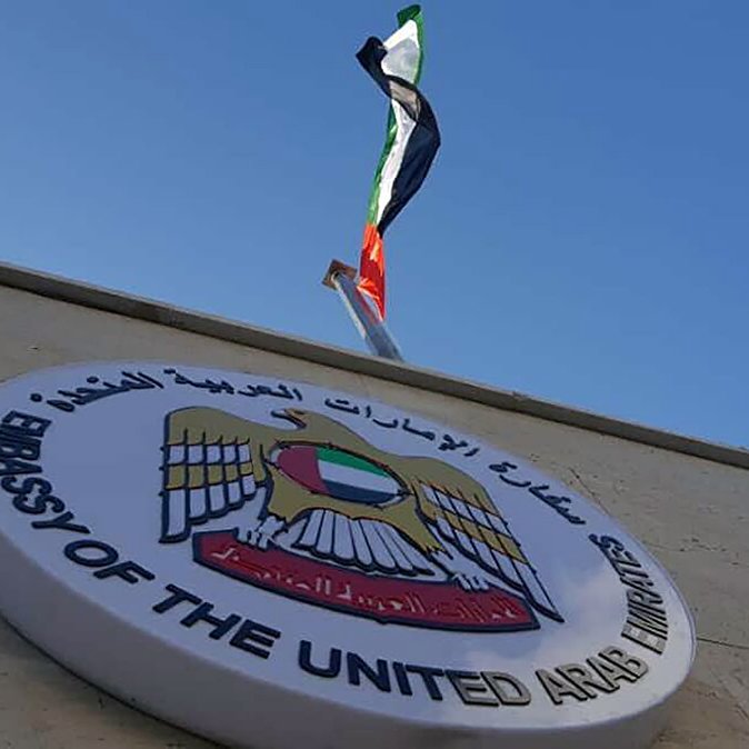 L'image montre un bâtiment avec un panneau qui indique qu'il s'agit de l'ambassade des Émirats arabes unis. Au-dessus du panneau, on peut voir le drapeau des Émirats arabes unis flottant. Le ciel est clair et légèrement nuageux, ce qui donne une belle arrière-plan à la scène.