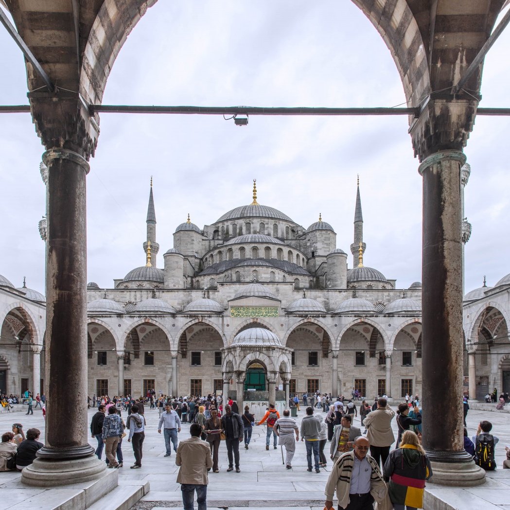 L'image montre une vue grandiose de la Mosquée bleue à Istanbul, encadrée par des arches majestueuses. Au premier plan, on peut voir un espace vaste et ouvert avec de nombreuses personnes qui se déplacent, créant une ambiance vivante. La mosquée elle-même se dresse en arrière-plan avec ses dômes et ses minarets élancés, sous un ciel nuageux qui ajoute une atmosphère particulière à la scène. Les détails architecturaux des colonnes et des arches sont également visibles, soulignant la beauté de cette œuvre historique.