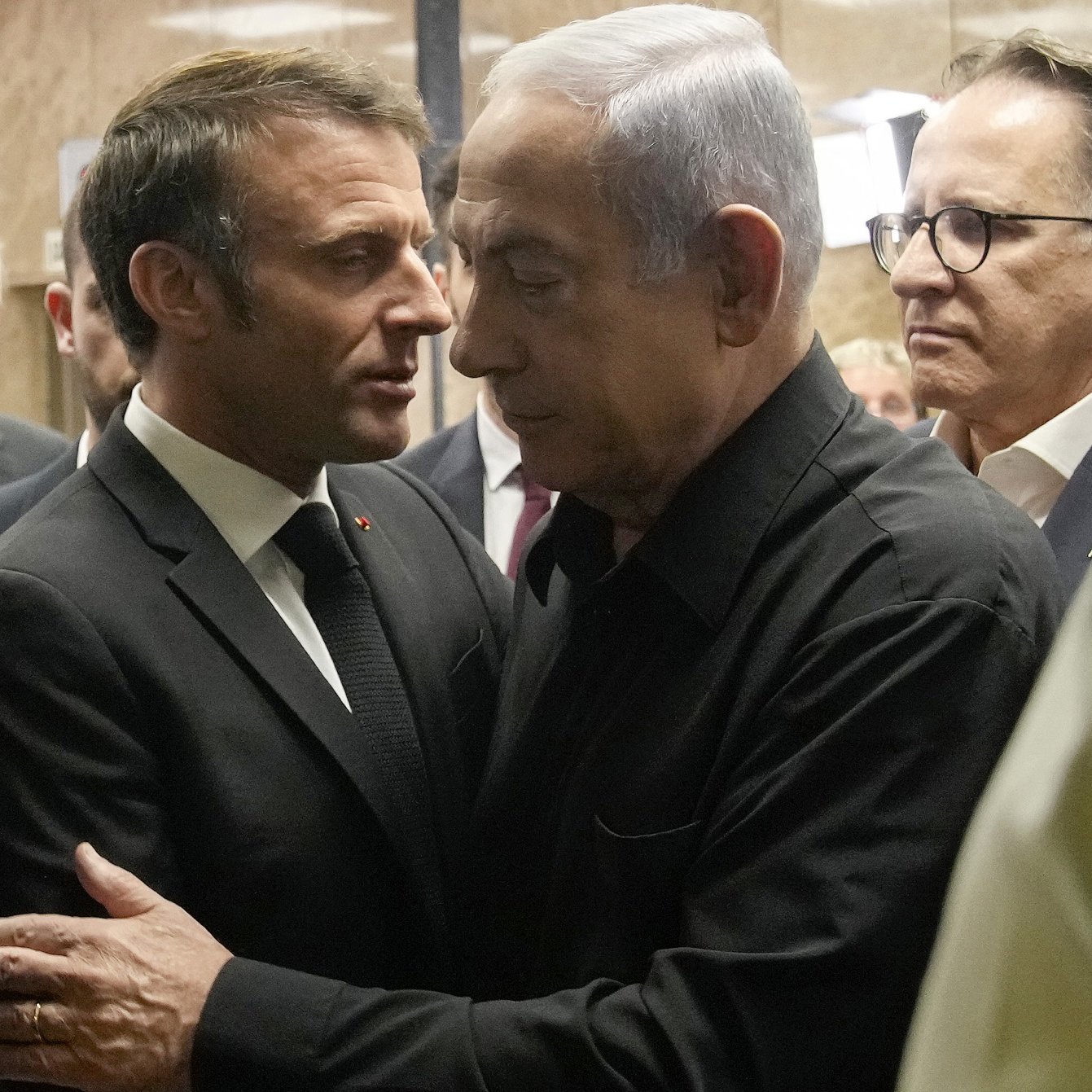 The image depicts two men in close proximity, engaged in a serious conversation. One man is dressed in a formal suit and tie, while the other is wearing a black shirt. They appear to be in an indoor setting, surrounded by several other individuals in the background, who are also dressed in formal attire. The atmosphere looks tense or important, suggesting a significant discussion is taking place.